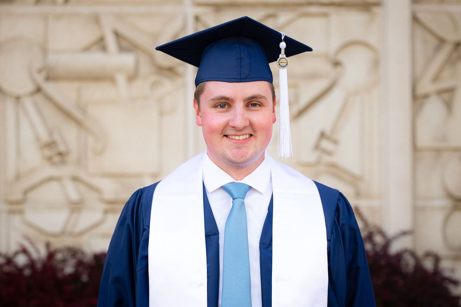 A smiling man in a graduation gown and cap stands in front of a brick building.