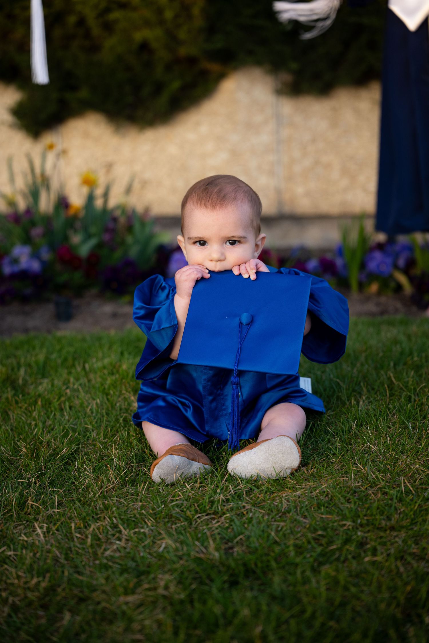 A baby boy sits on the grass in a blue outfit, looking at the camera.