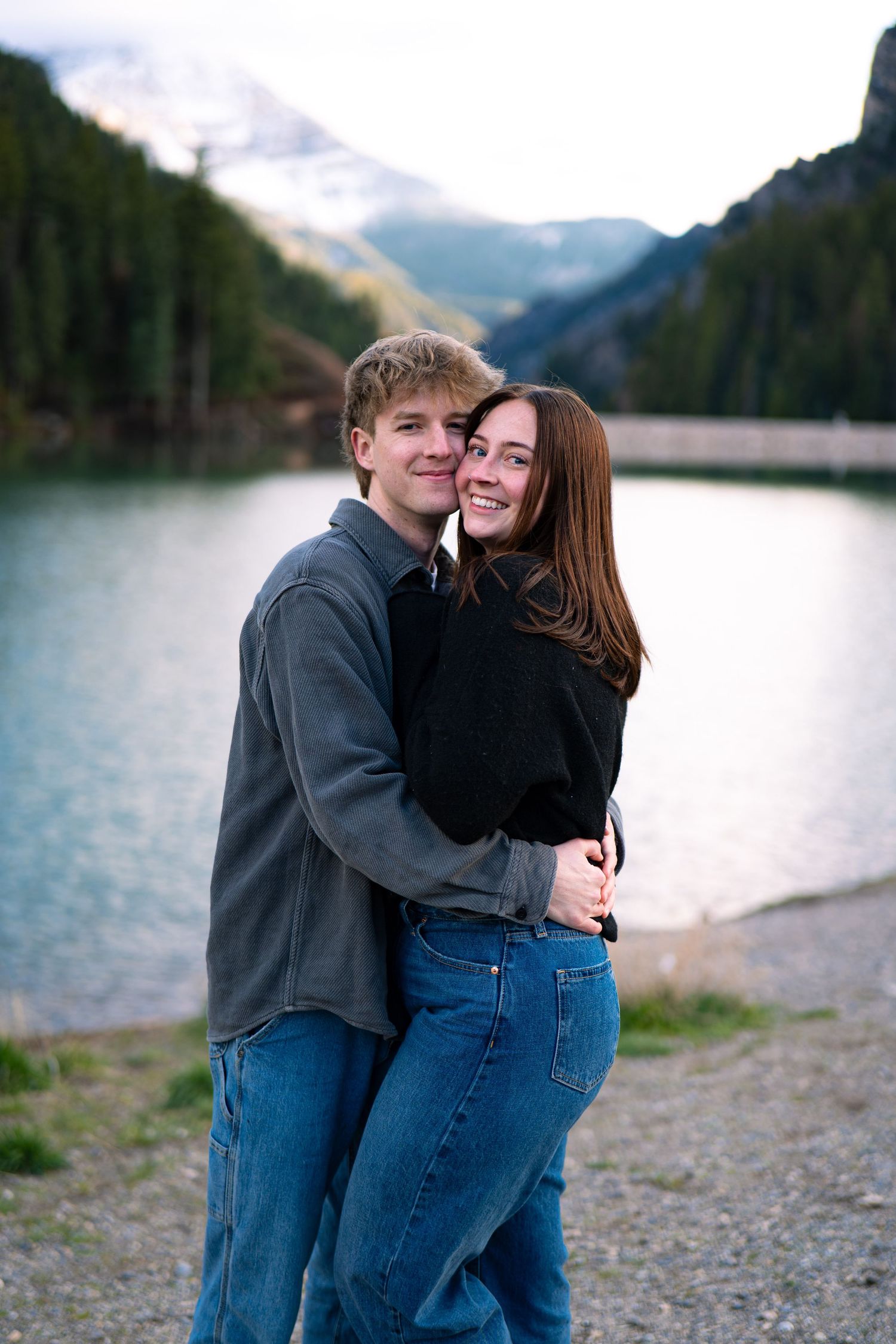 Young couple embracing and smiling by a mountain lake with snow-capped peaks and forest backdrop.