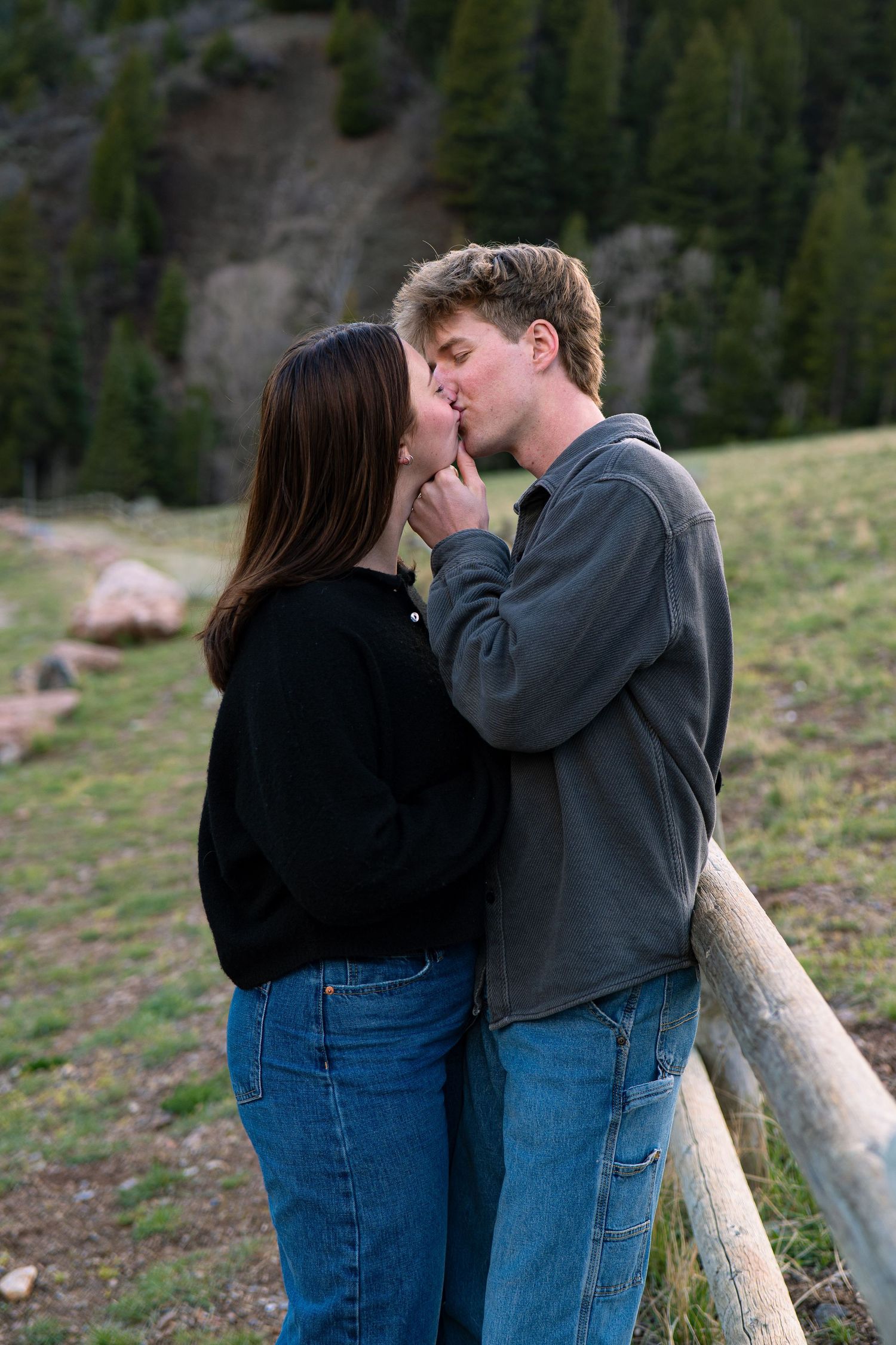 Young couple sharing a romantic kiss outdoors near a wooden fence with green mountain scenery in the background.