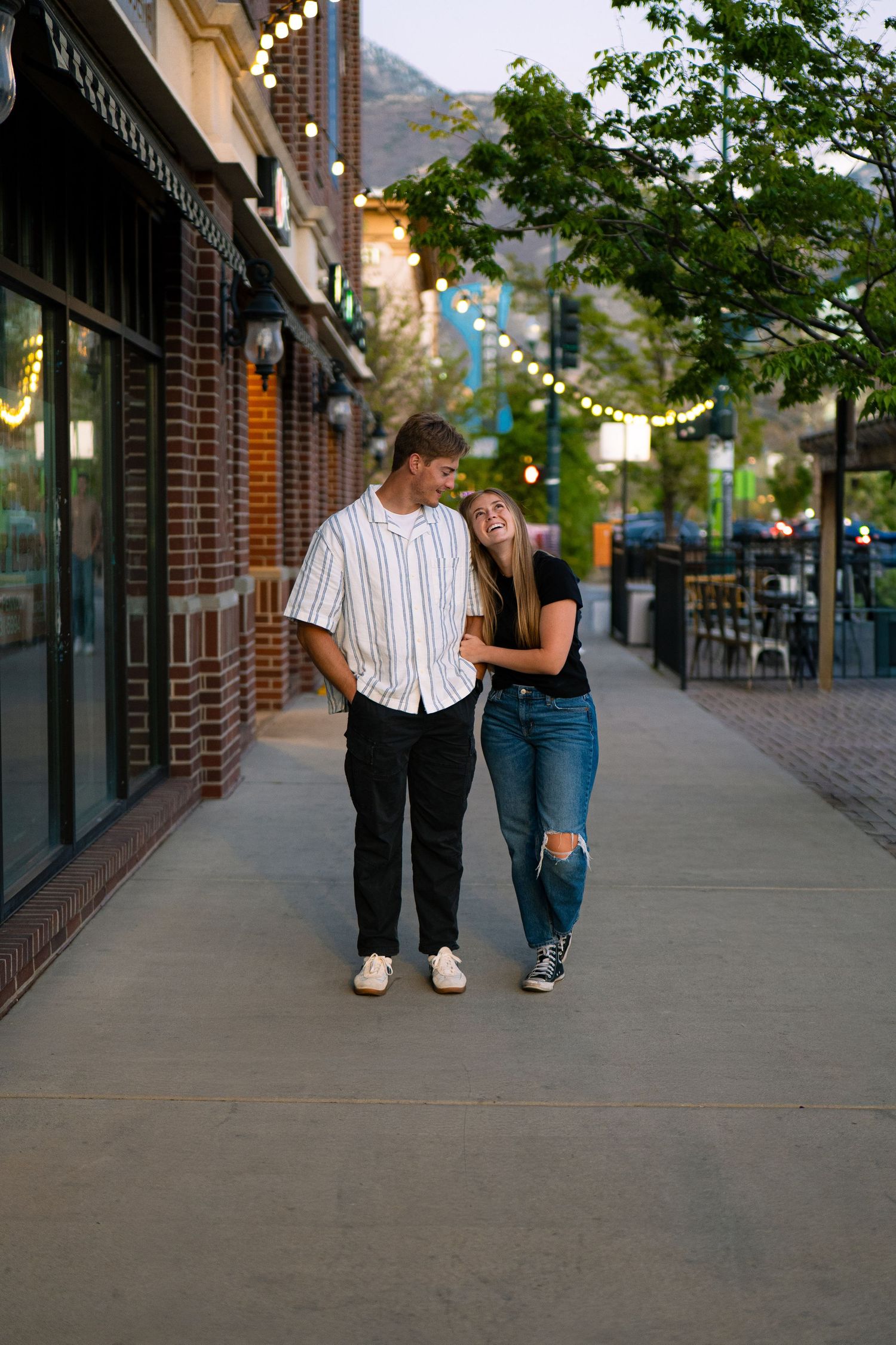 A happy couple walks arm-in-arm along a charming urban sidewalk with string lights and brick buildings at dusk.