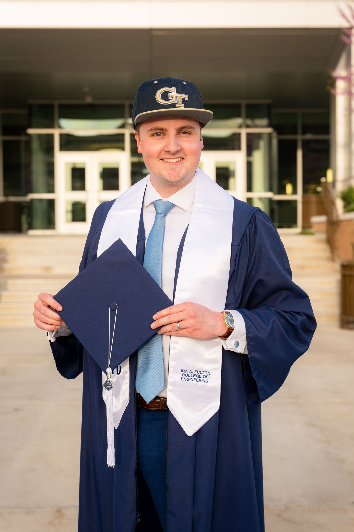 A man in a graduation gown and cap stands in front of a university building.