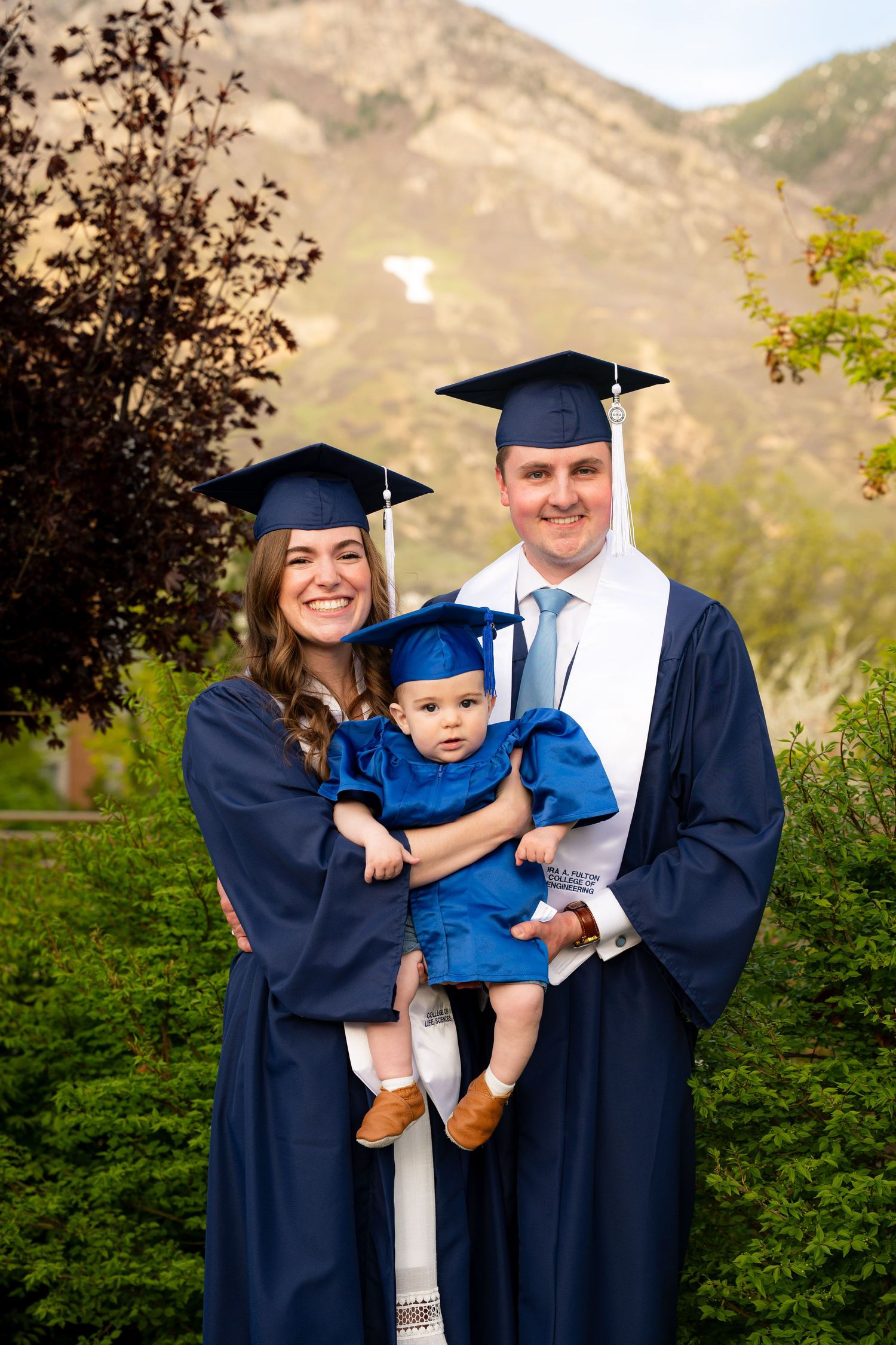A family of three, including a baby, pose together in their graduation gowns in a scenic outdoor setting.