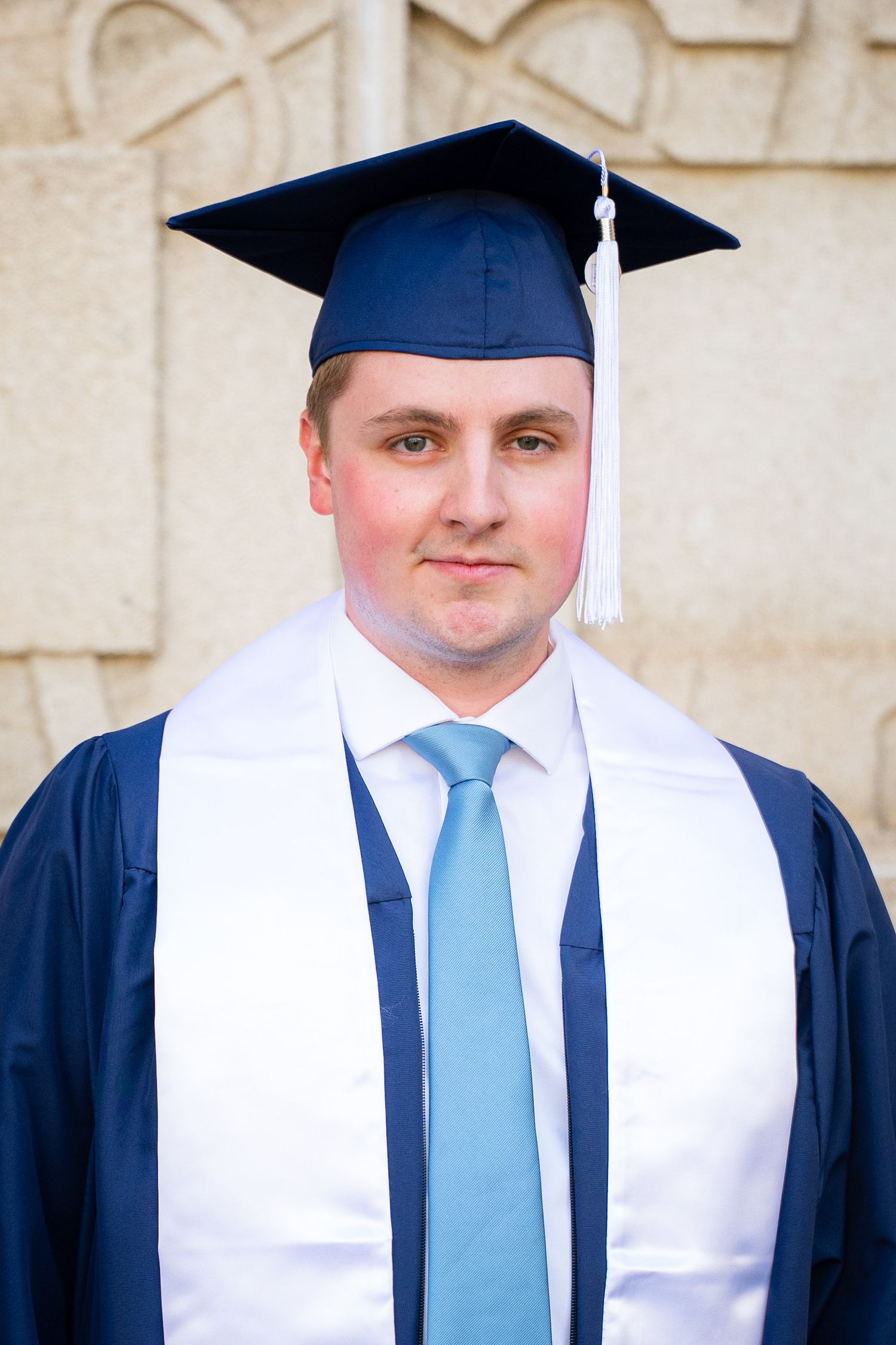 A smiling man in a graduation gown and cap stands in front of a brick building.