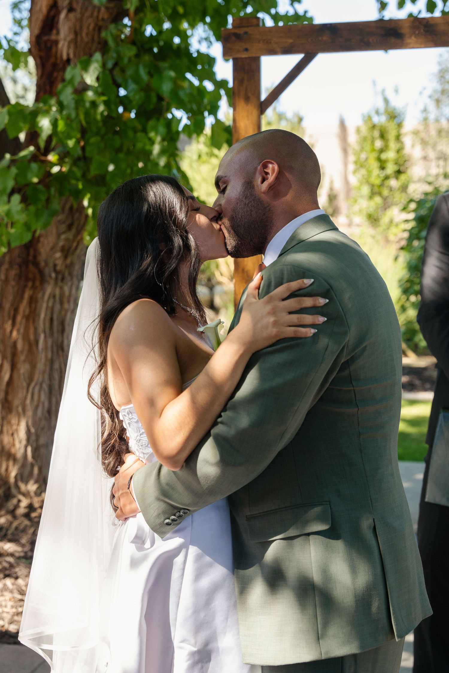 Bride and groom share their first kiss at outdoor wedding ceremony under wooden arbor surrounded by green trees.