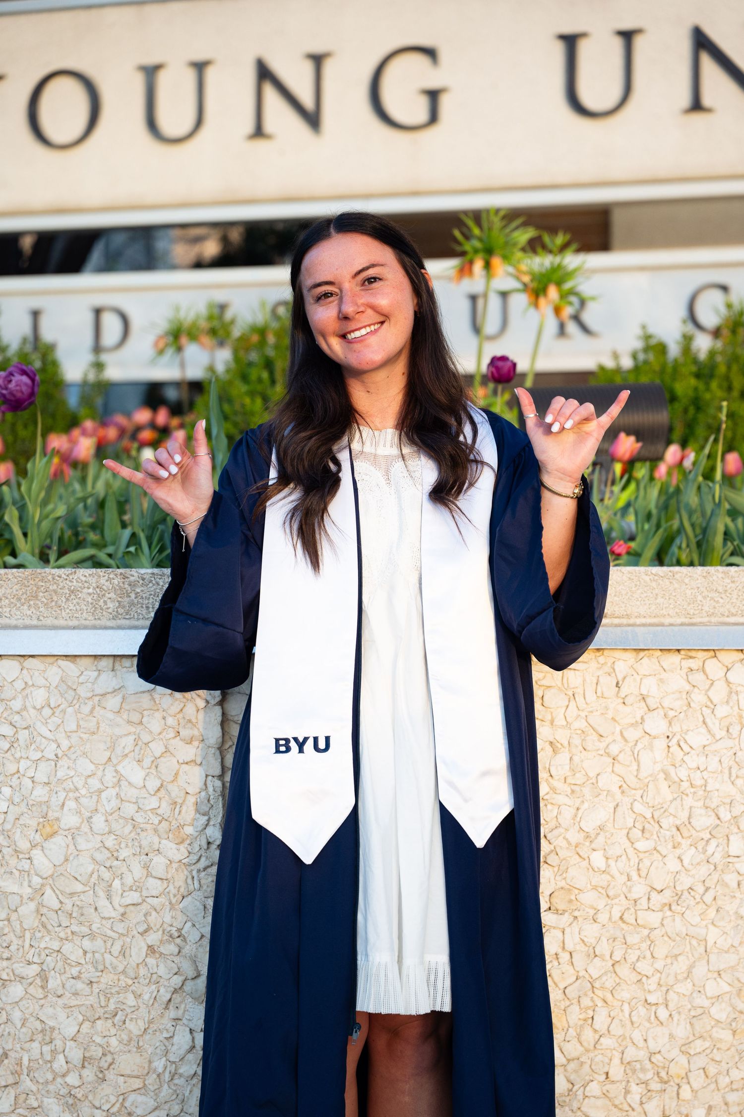 BYU graduate posing with shaka signs in navy gown and white BYU stole outside Brigham Young University.