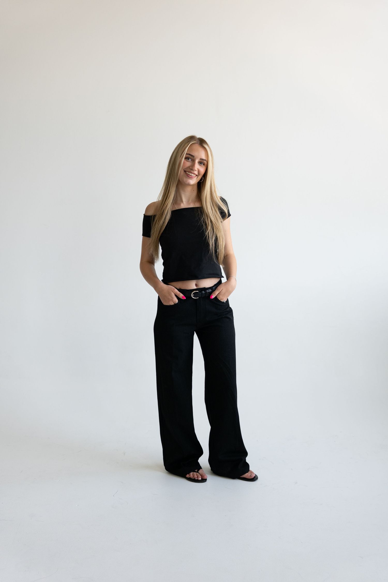 Young woman wearing black crop top, wide-leg pants, and belt, posing confidently against white background.