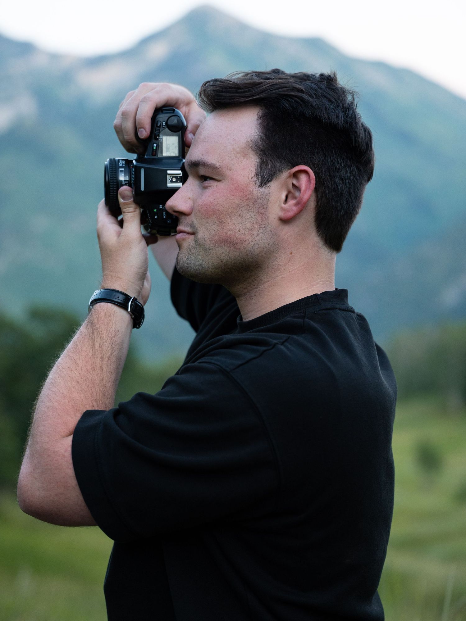 Man in black shirt photographing mountain landscape with DSLR camera outdoors.