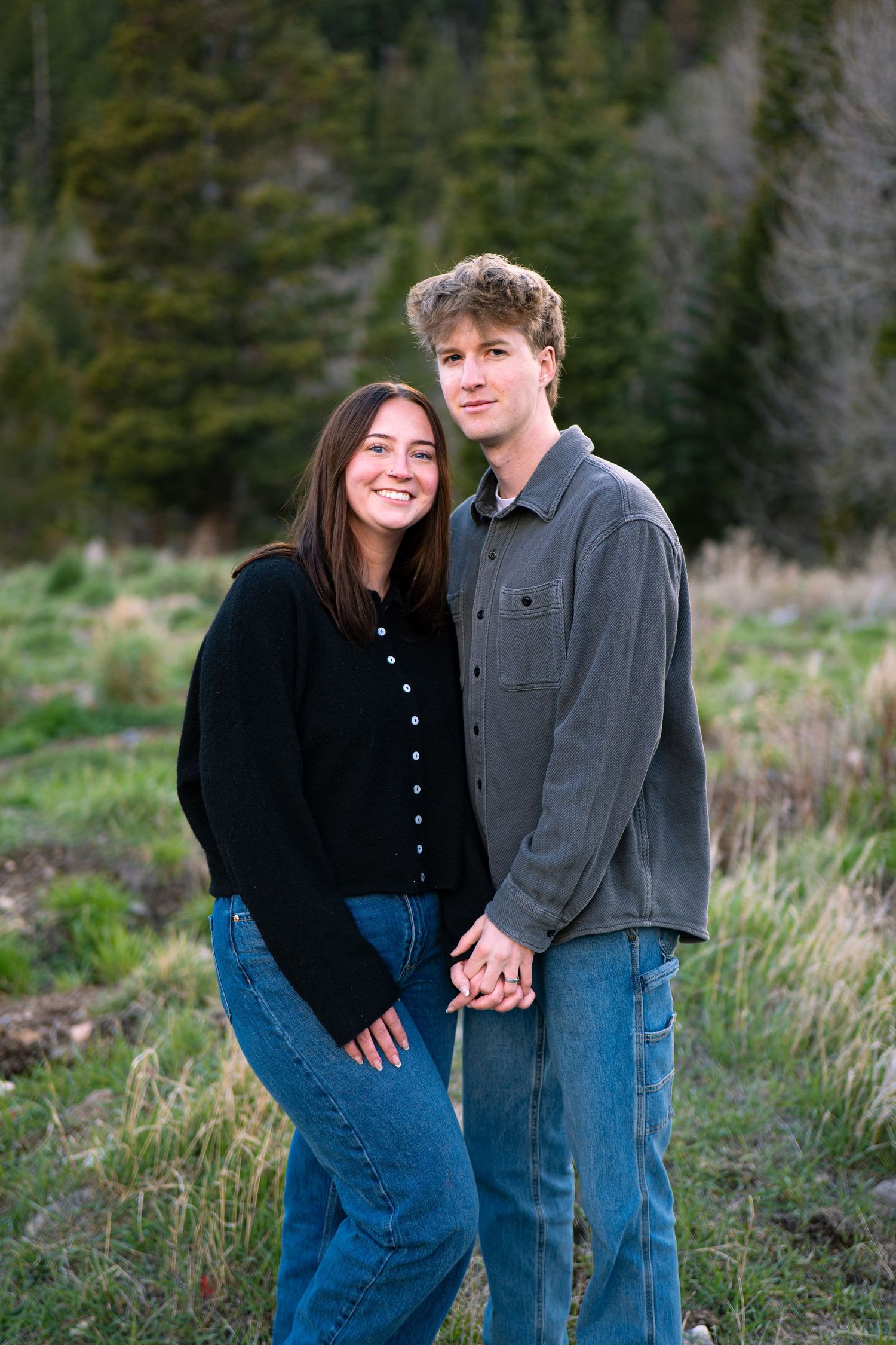 A young couple holding hands and smiling outdoors in a mountain meadow with evergreen trees in the background.