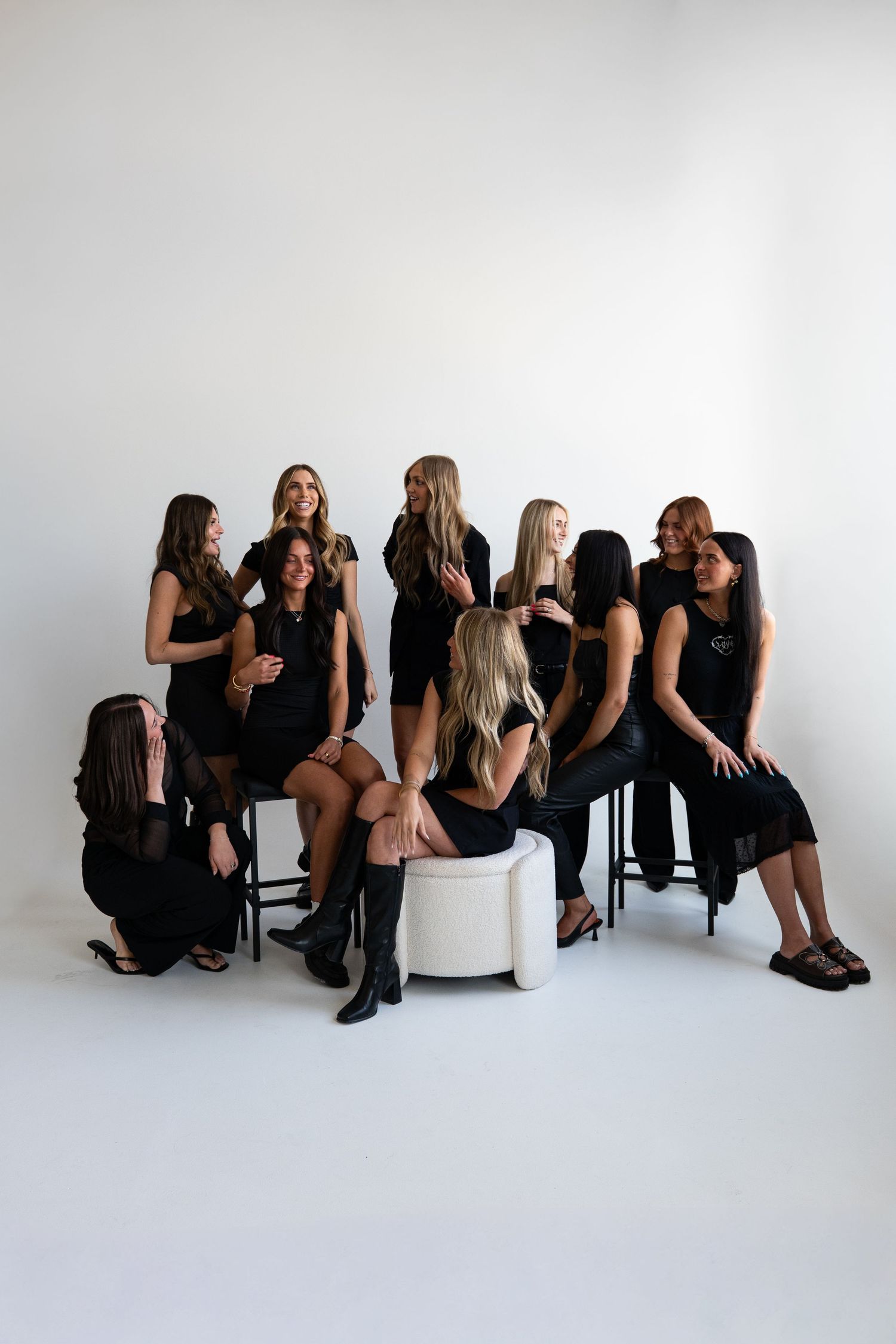 Group of nine women in matching black outfits posing together in a bright white studio setting.