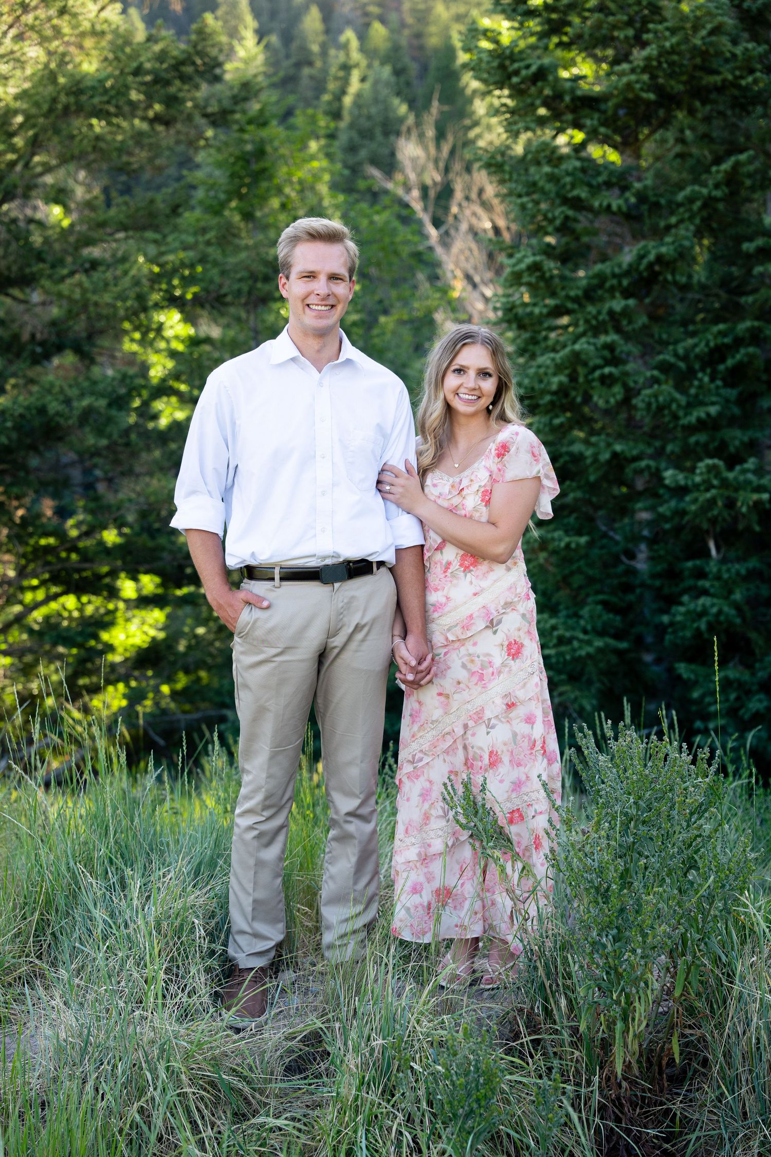 A smiling couple poses outdoors in lush green surroundings, man in white shirt and woman in floral dress.