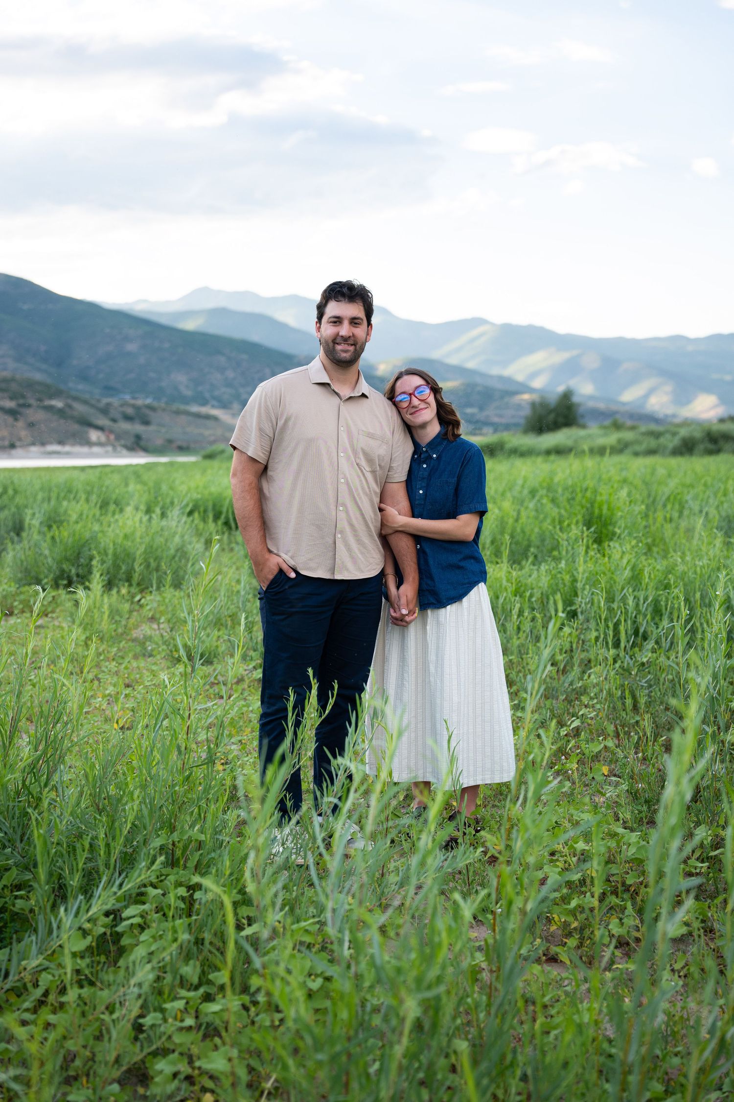 A couple stands smiling in a lush green mountain meadow, the woman leaning on the man's shoulder.