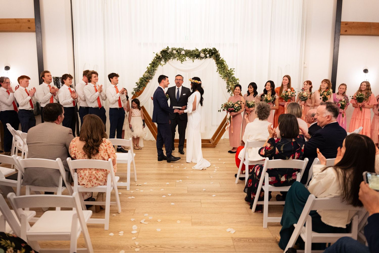 Indoor wedding ceremony with couple at floral arch altar, bridesmaids in pink, groomsmen in white, guests seated on white chairs.