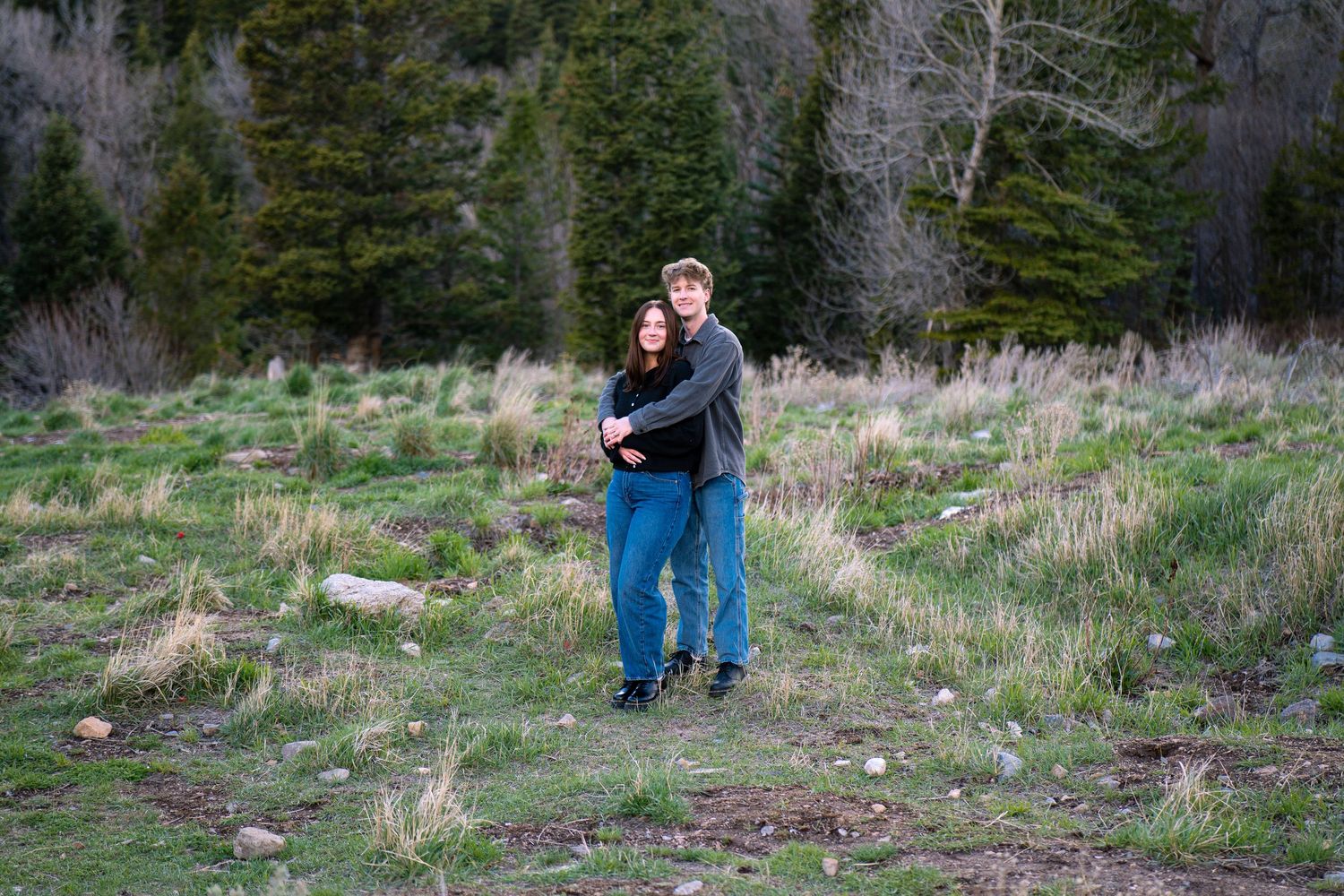 Couple embracing in a lush green meadow surrounded by evergreen trees during an outdoor engagement photo session.