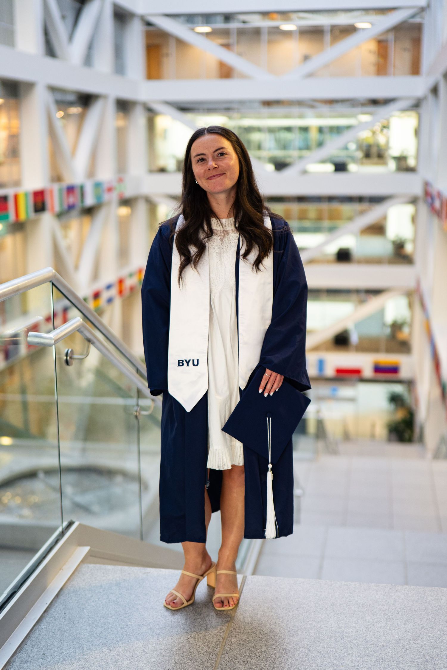 Woman in navy BYU graduation gown and white stole poses on stairs in modern university building.