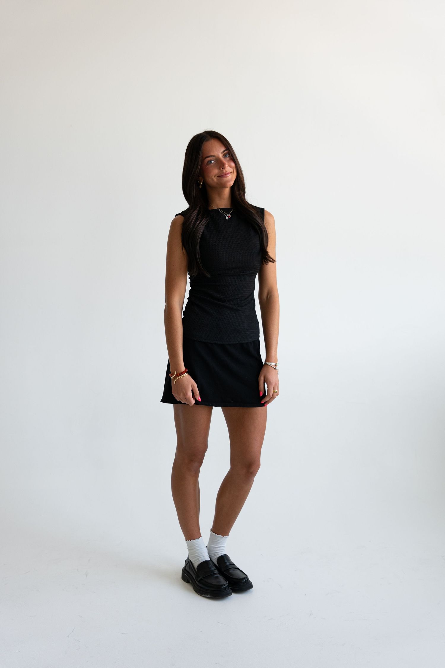 Woman in black sleeveless top and mini skirt with white socks and loafers on white background.