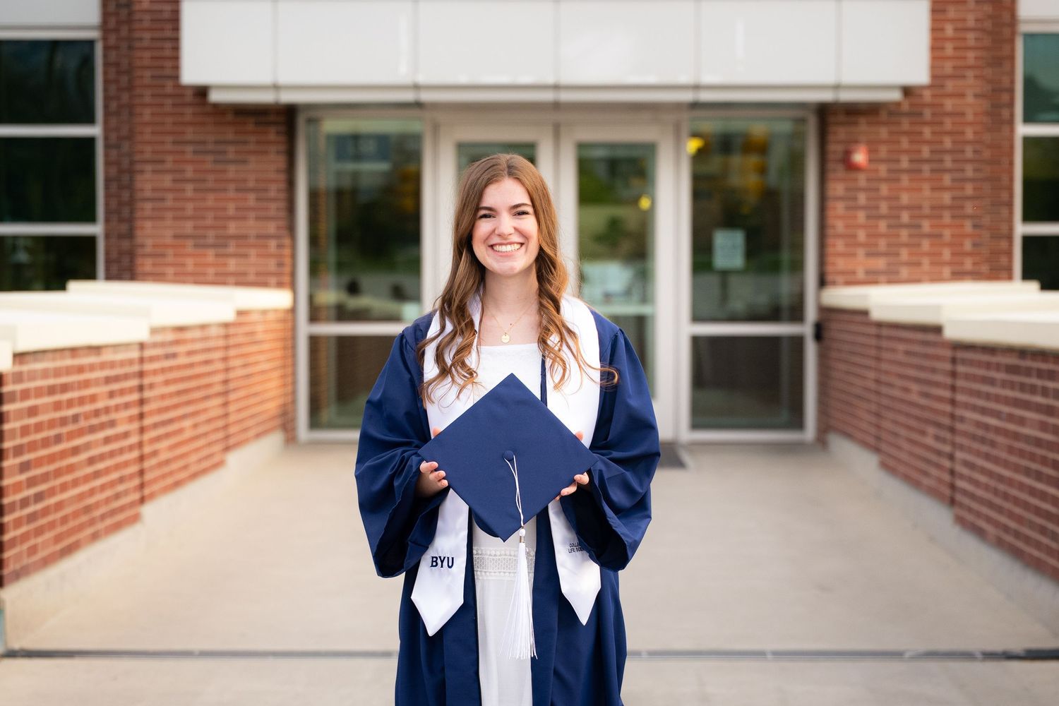 A young woman with long curly brown hair smiles while wearing a white dress and blue graduation gown in front of a building.