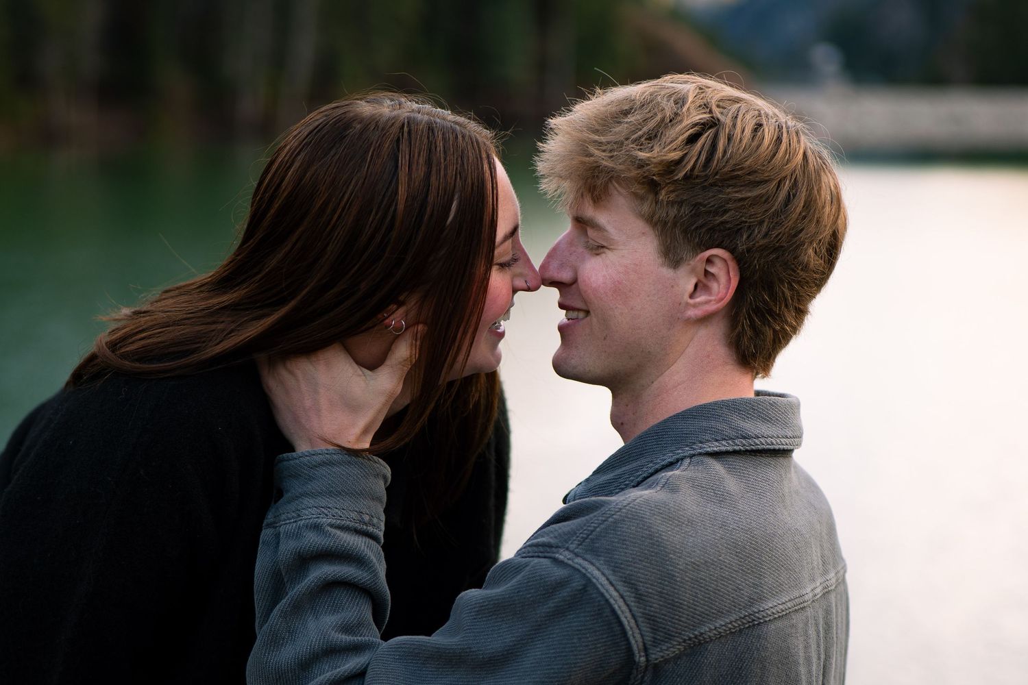 A young couple shares a romantic moment by a lake, smiling and about to kiss outdoors.