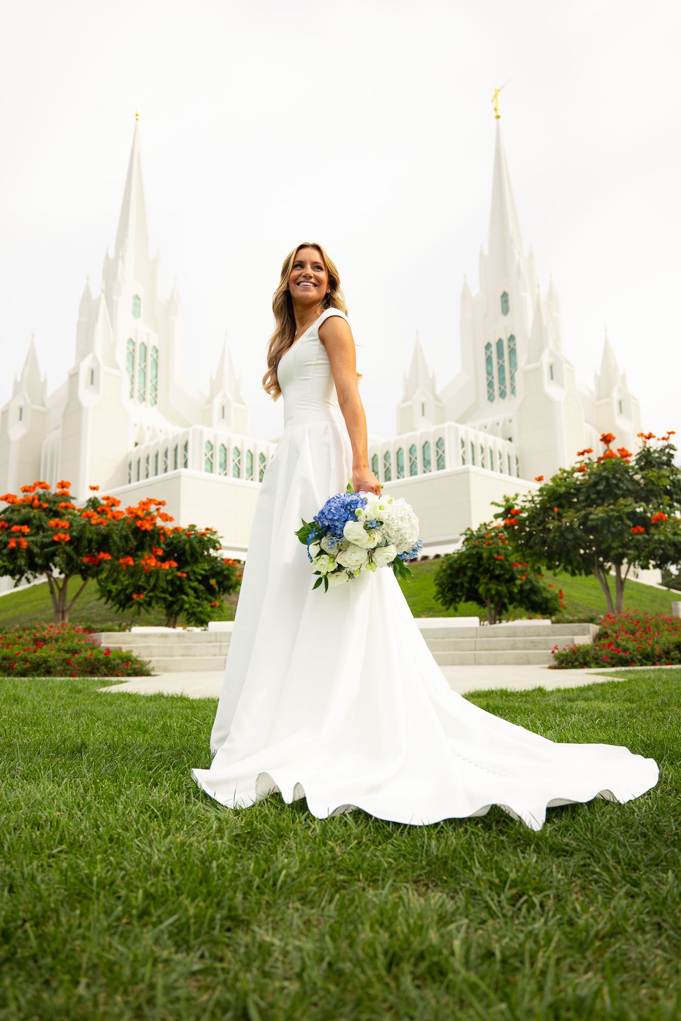 Bride in white gown holding blue and white bouquet stands before a white LDS temple with spires and orange flowers.