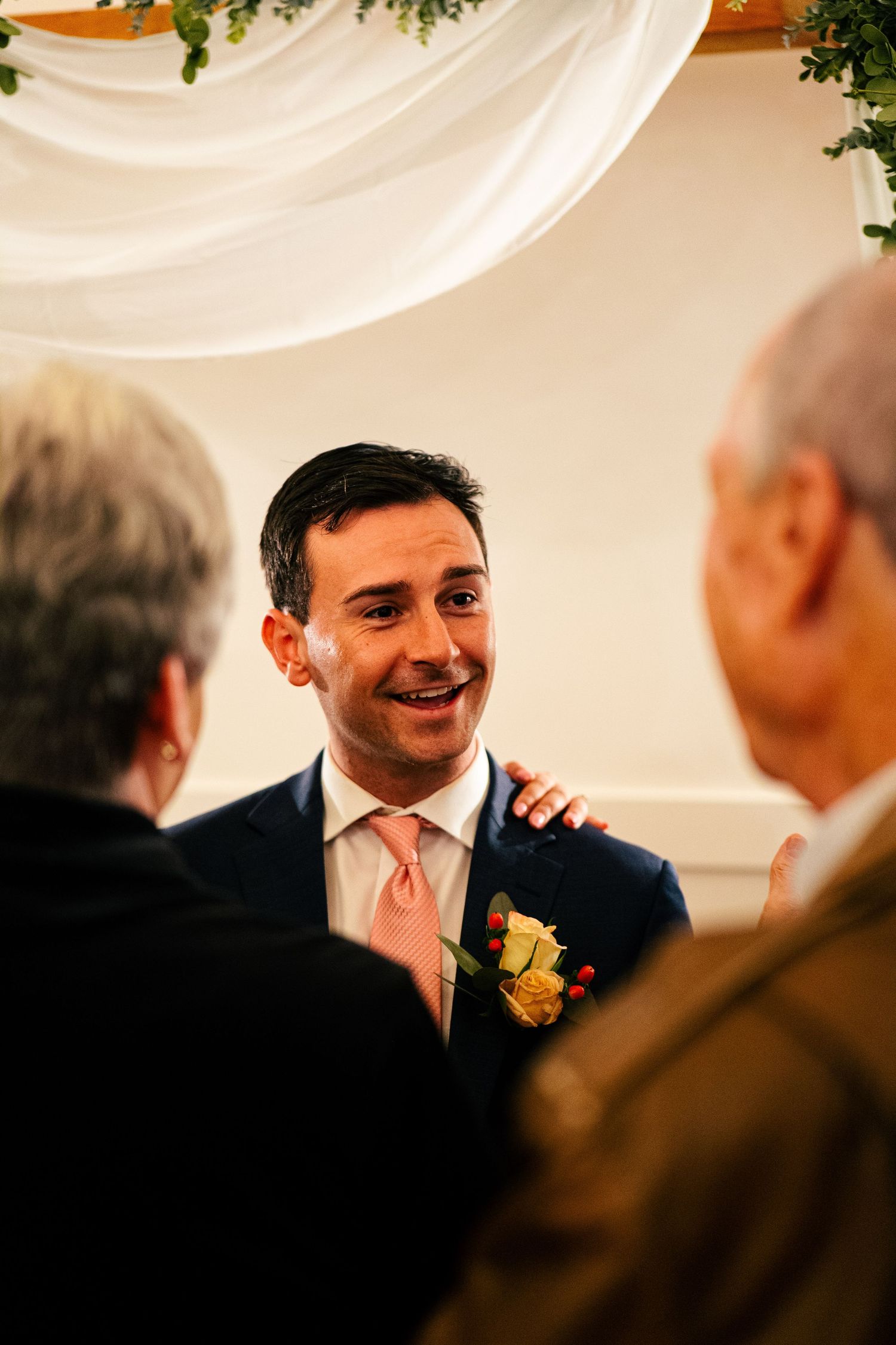 Smiling groom in navy suit with yellow boutonniere greeting guests at indoor wedding ceremony.