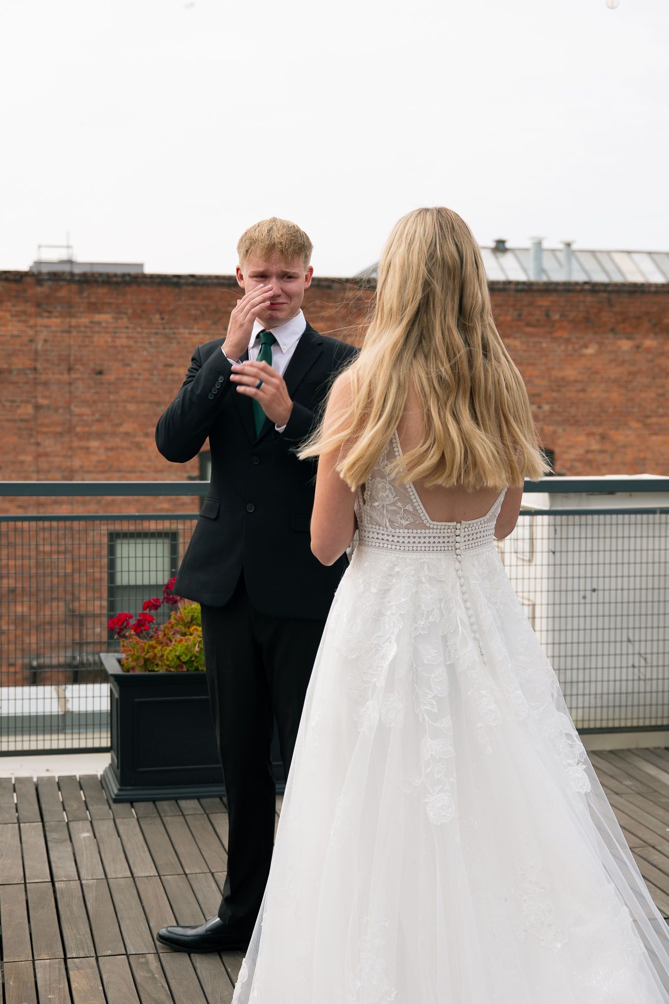 Emotional groom wiping tears seeing bride in white lace wedding gown during first look on rooftop venue.