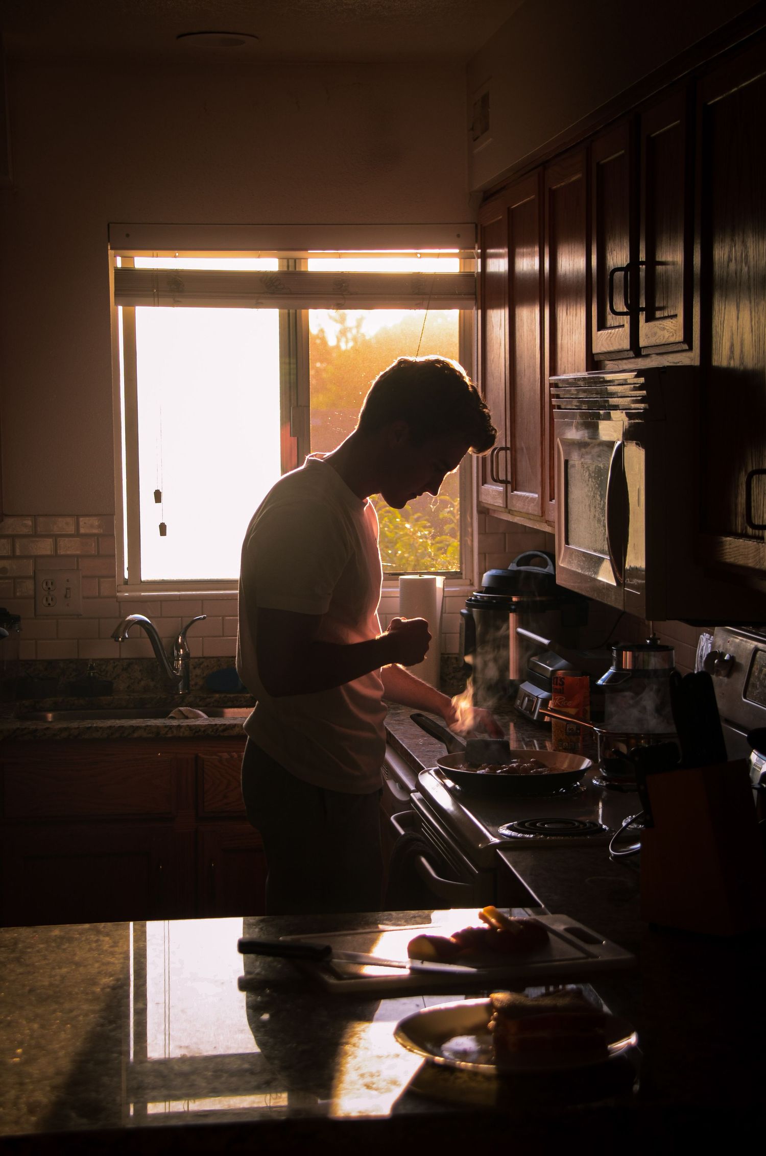 A silhouetted figure standing in a kitchen, preparing food near a window with the sunset visible.