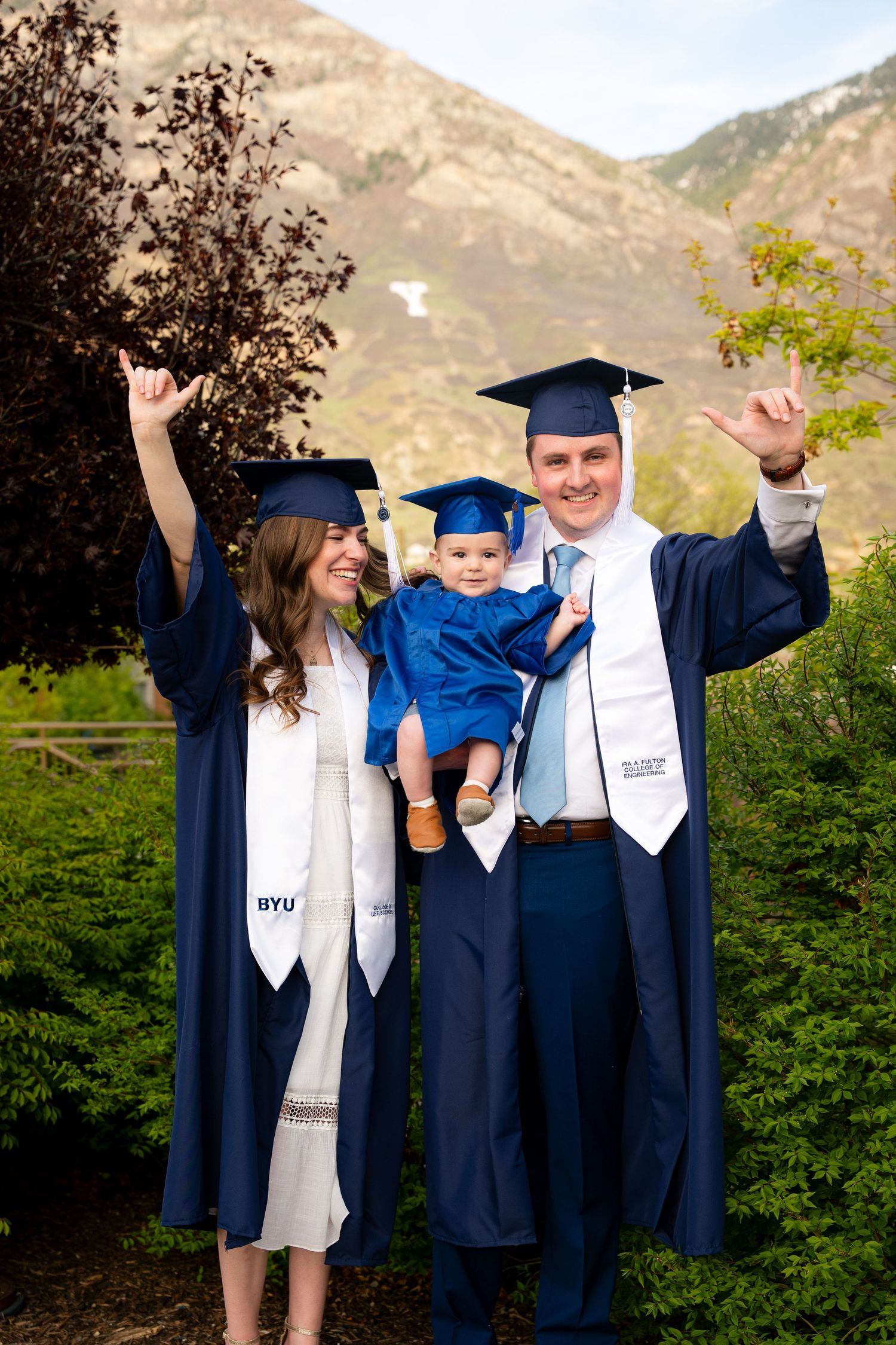 A family of three, including a baby, proudly pose together in their graduation gowns in a scenic outdoor setting.