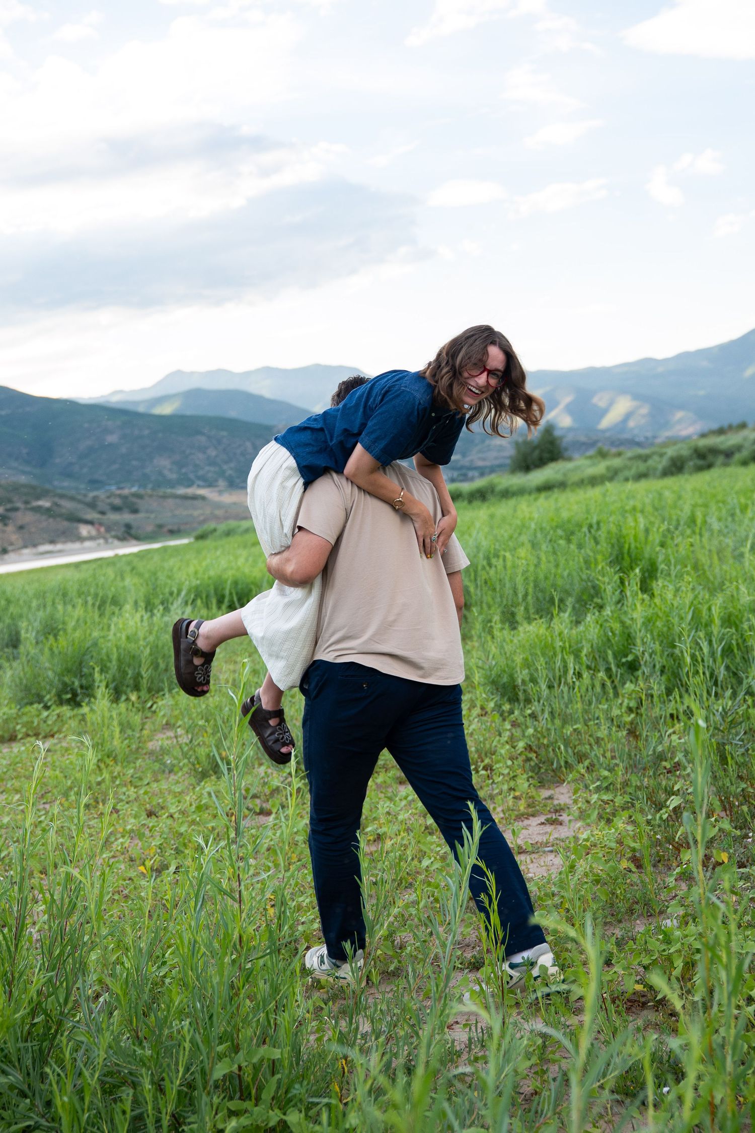 Couple playing in green meadow, man lifting woman on his shoulders with mountain backdrop.