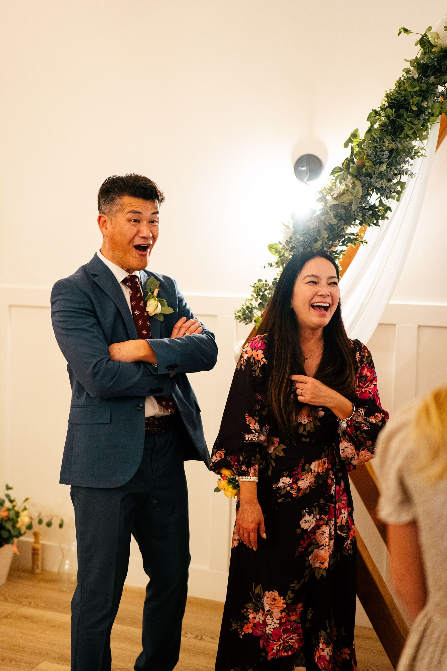 Man in navy suit and woman in floral dress laughing joyfully at an elegant indoor wedding reception.