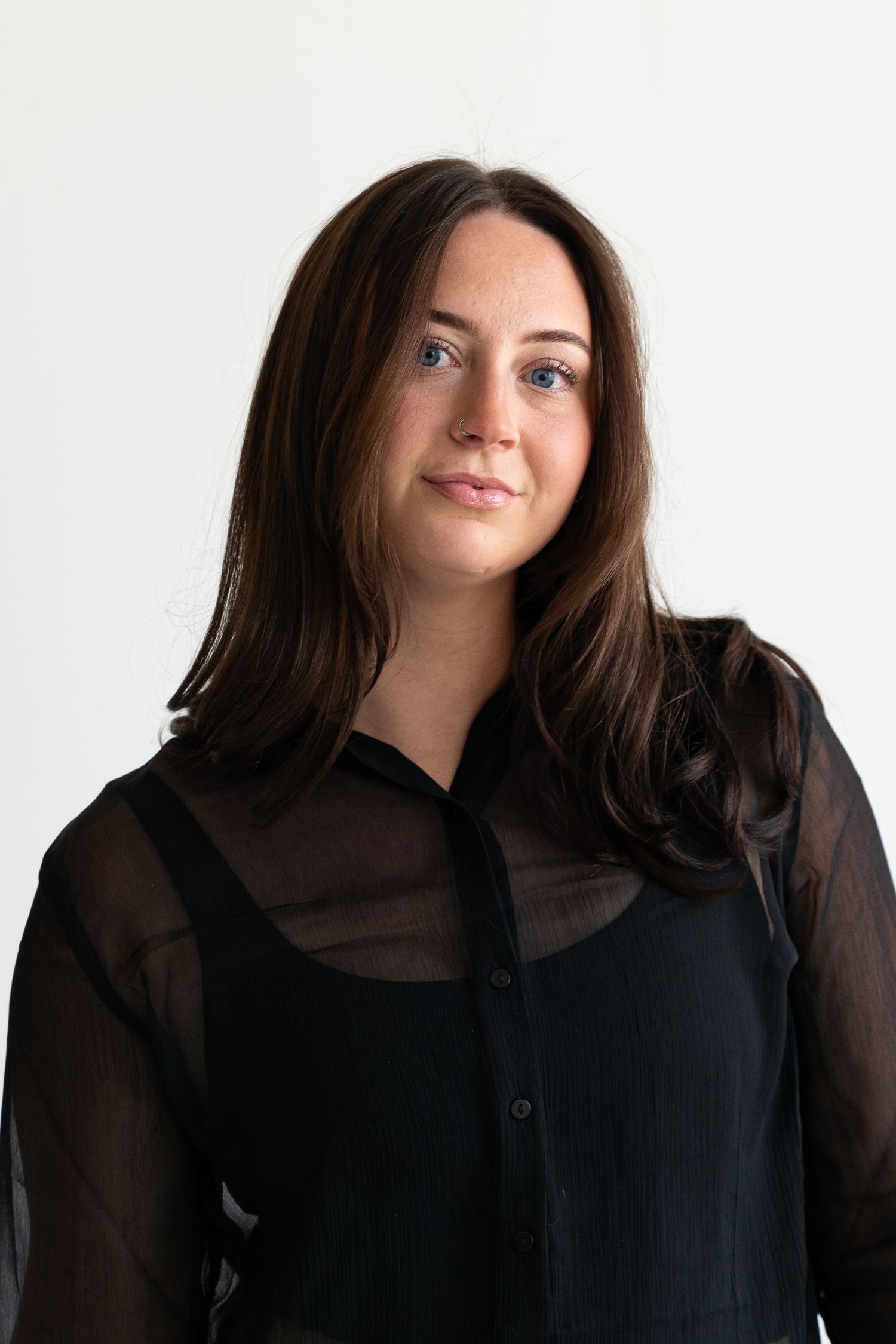 Young woman with long brown hair and blue eyes wearing a black sheer blouse, posing against a white background.