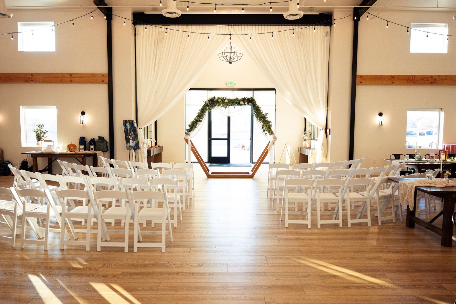 Elegant wedding venue with white chairs, hexagonal arch, greenery, string lights, and sheer drapes on hardwood floors.