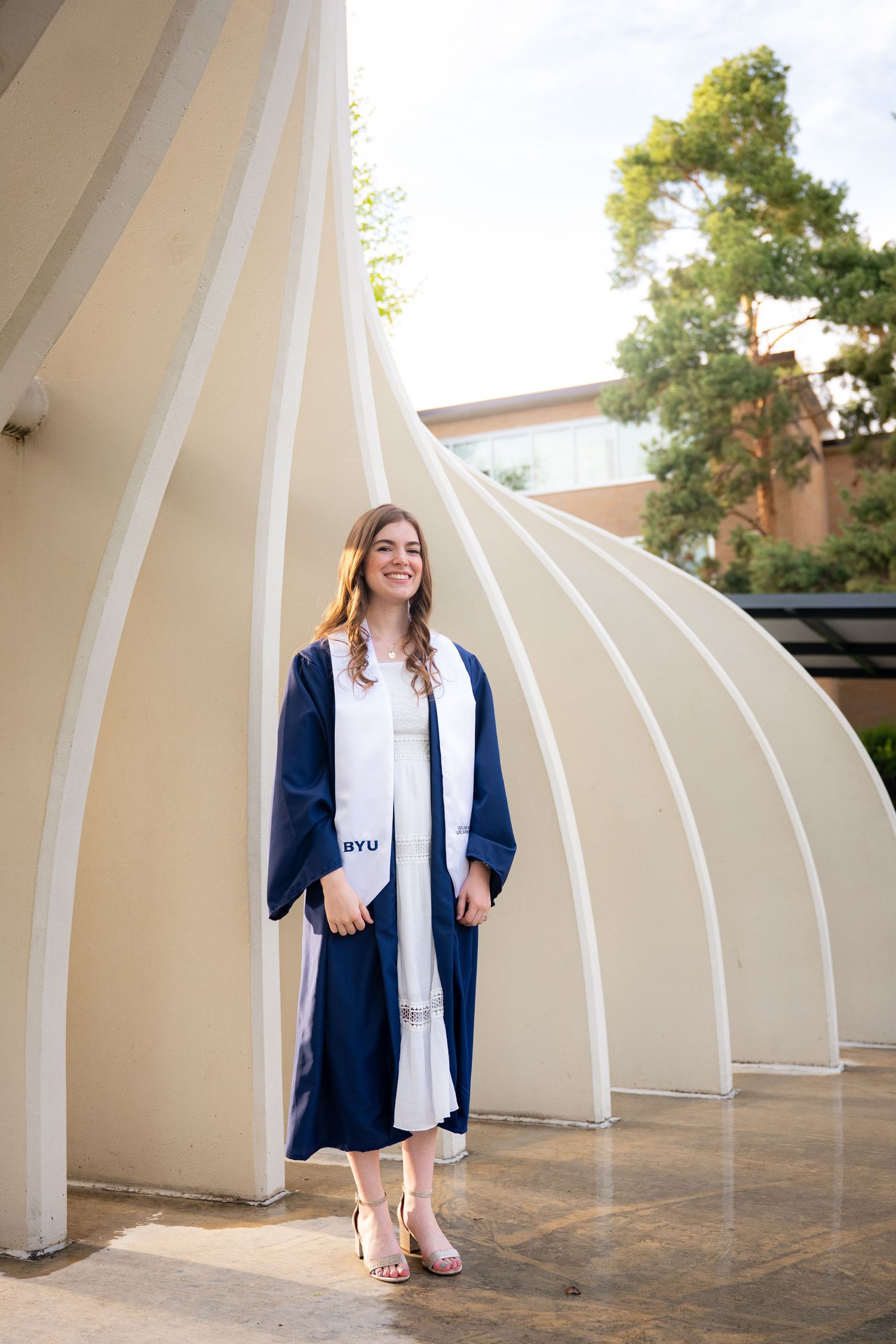 A young woman in a graduation gown stands in front of an architectural structure, smiling.