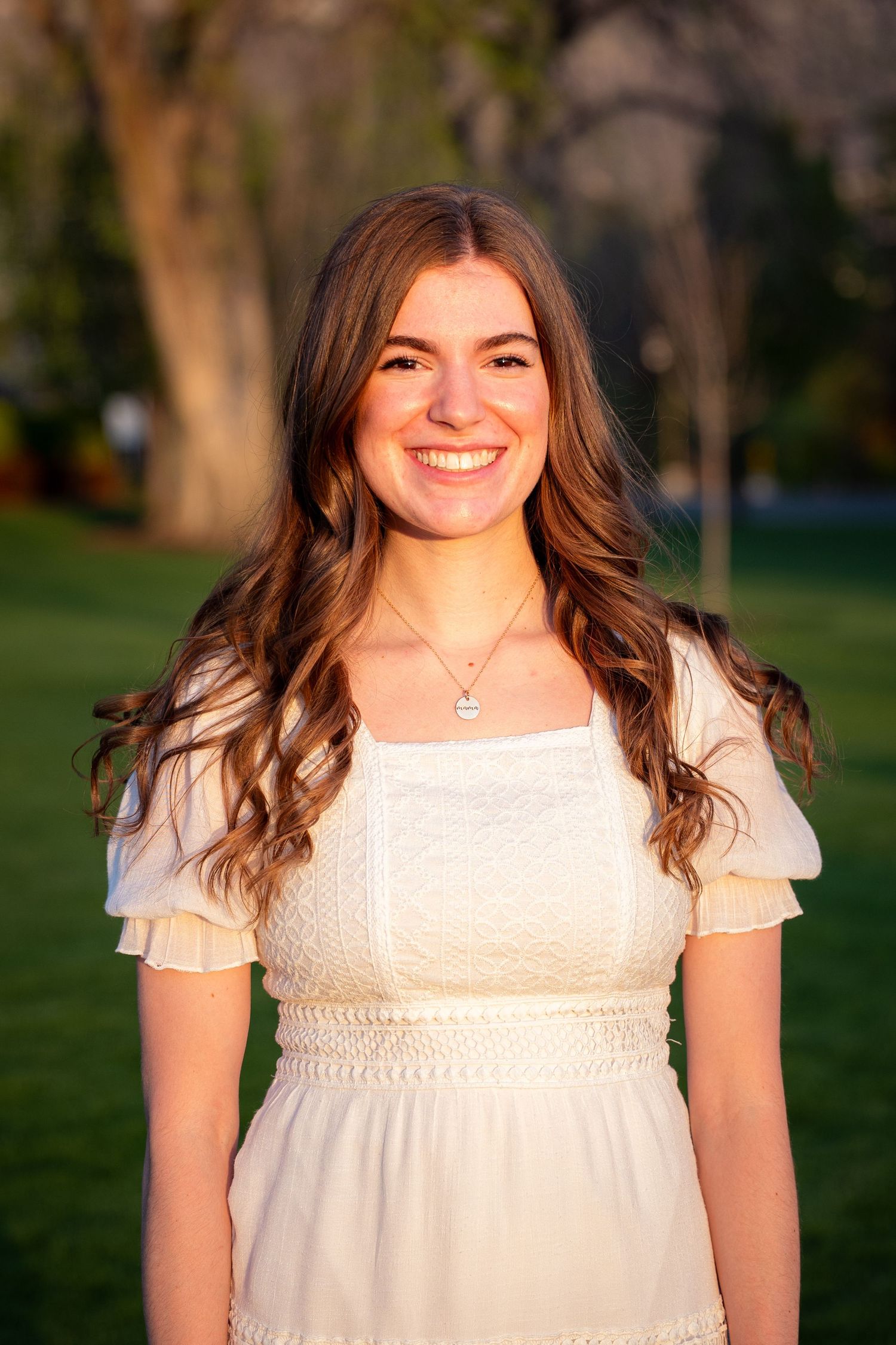 A young woman with long curly brown hair smiles while wearing a white dress in a grassy outdoor setting.