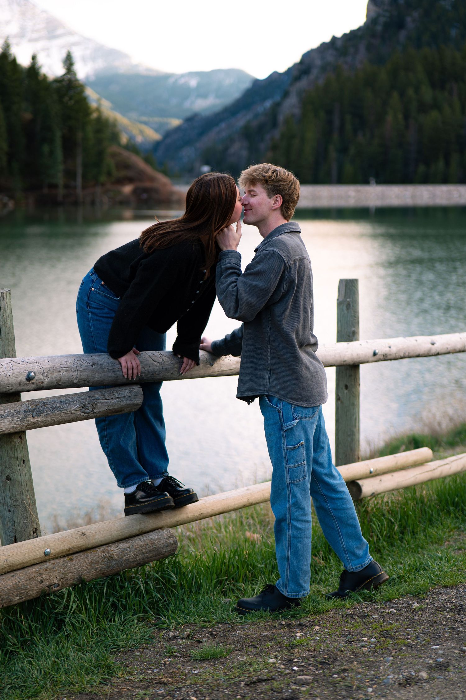 Young couple sharing a romantic moment by a rustic wooden fence with a mountain lake and forest backdrop.