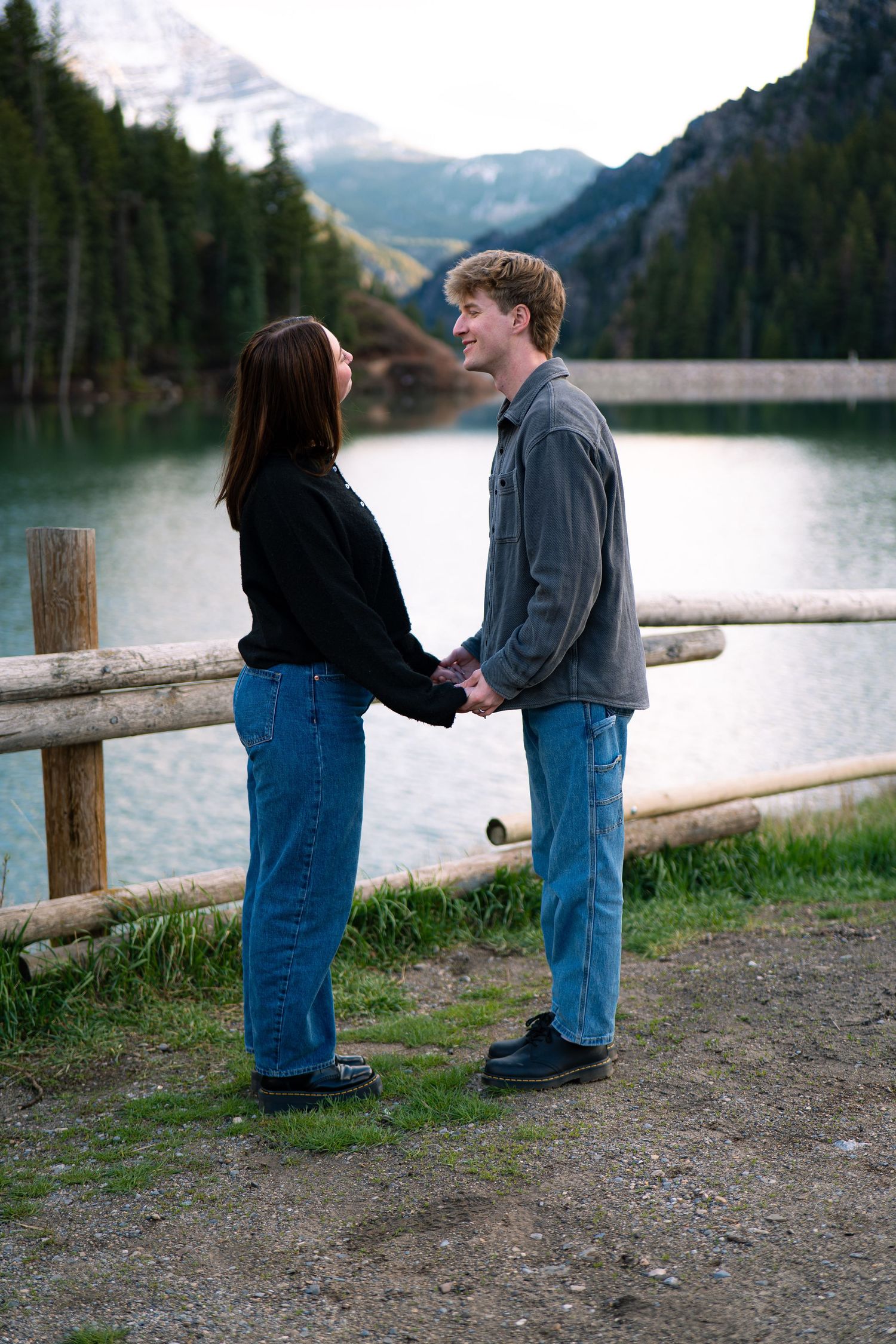 Young couple holding hands by a mountain lake with wooden fence and pine trees in background.