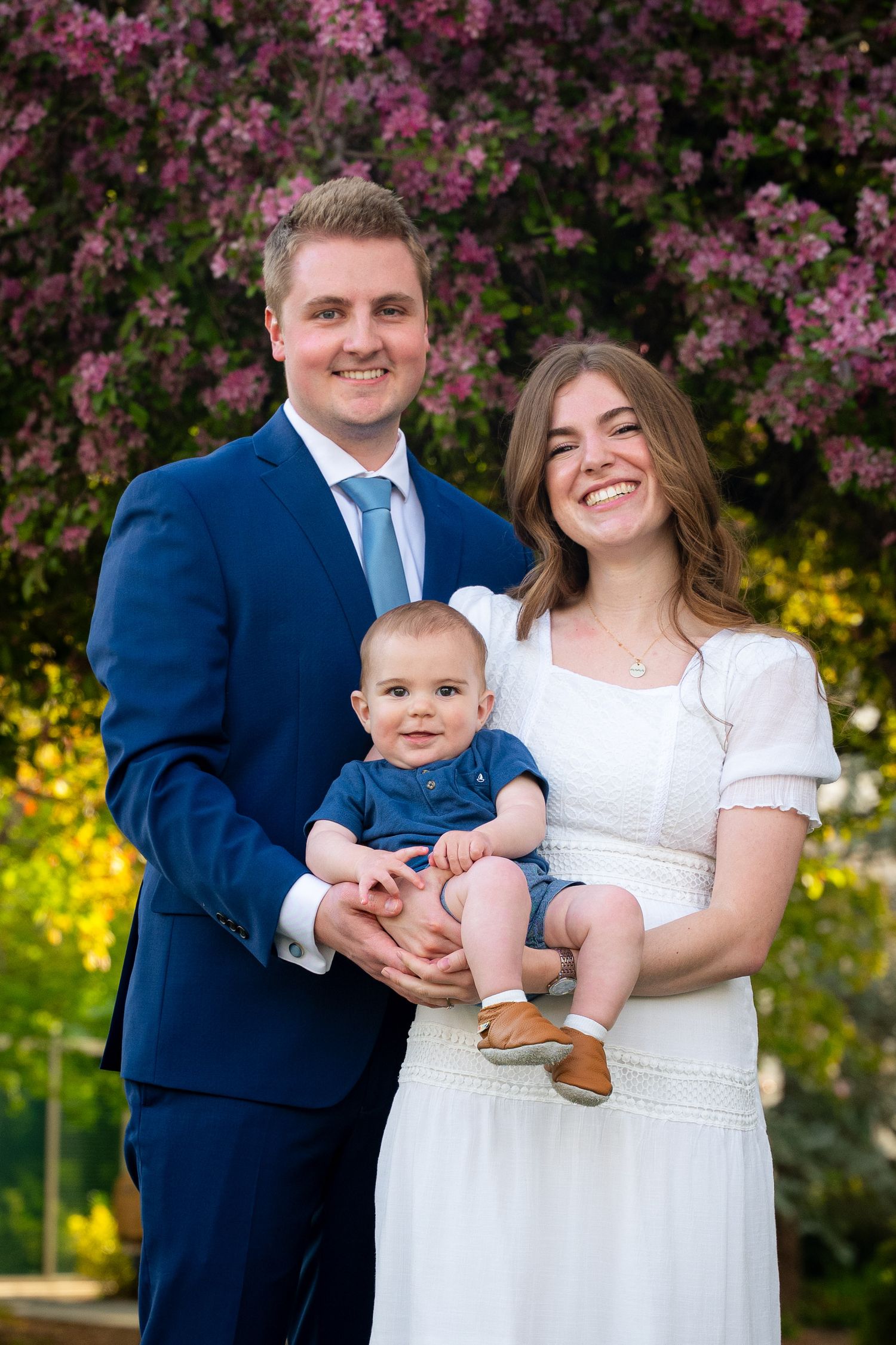 A family portrait of a couple holding a baby outdoors with flowers in the background.