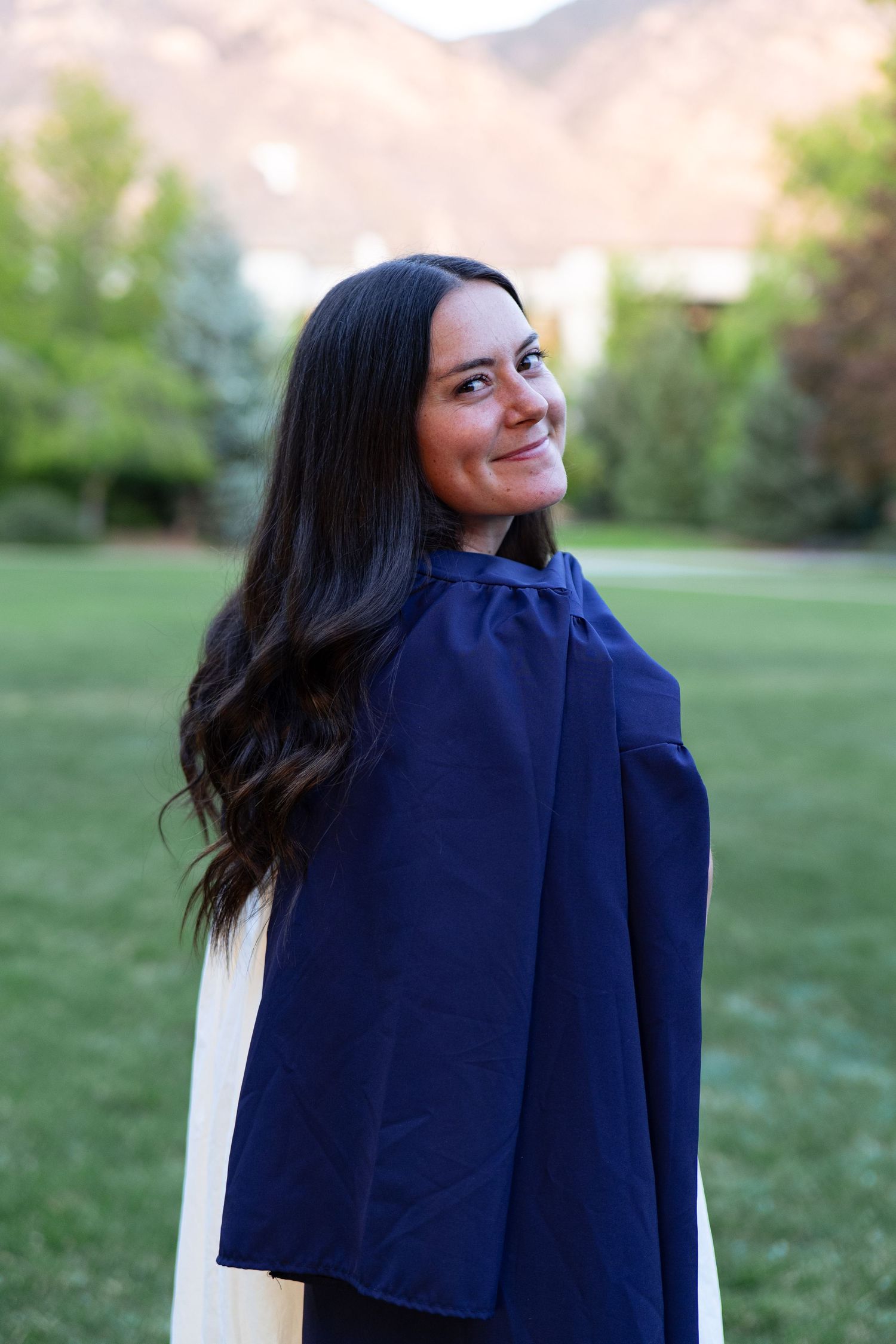Smiling female BYU graduate in navy commencement gown outdoors with mountain backdrop.