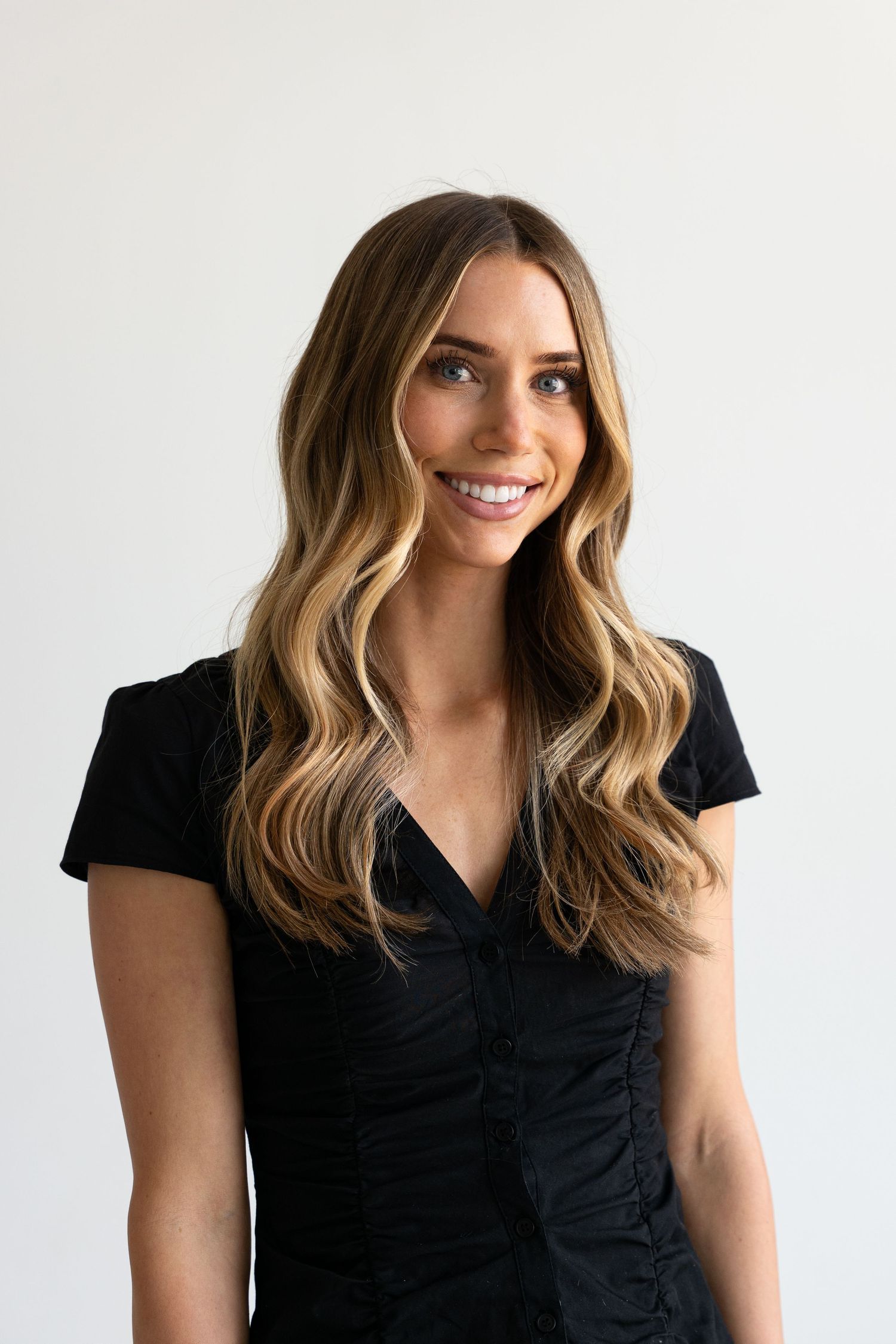 Smiling woman with long wavy blonde highlighted hair wearing a black ruched V-neck top against white background.