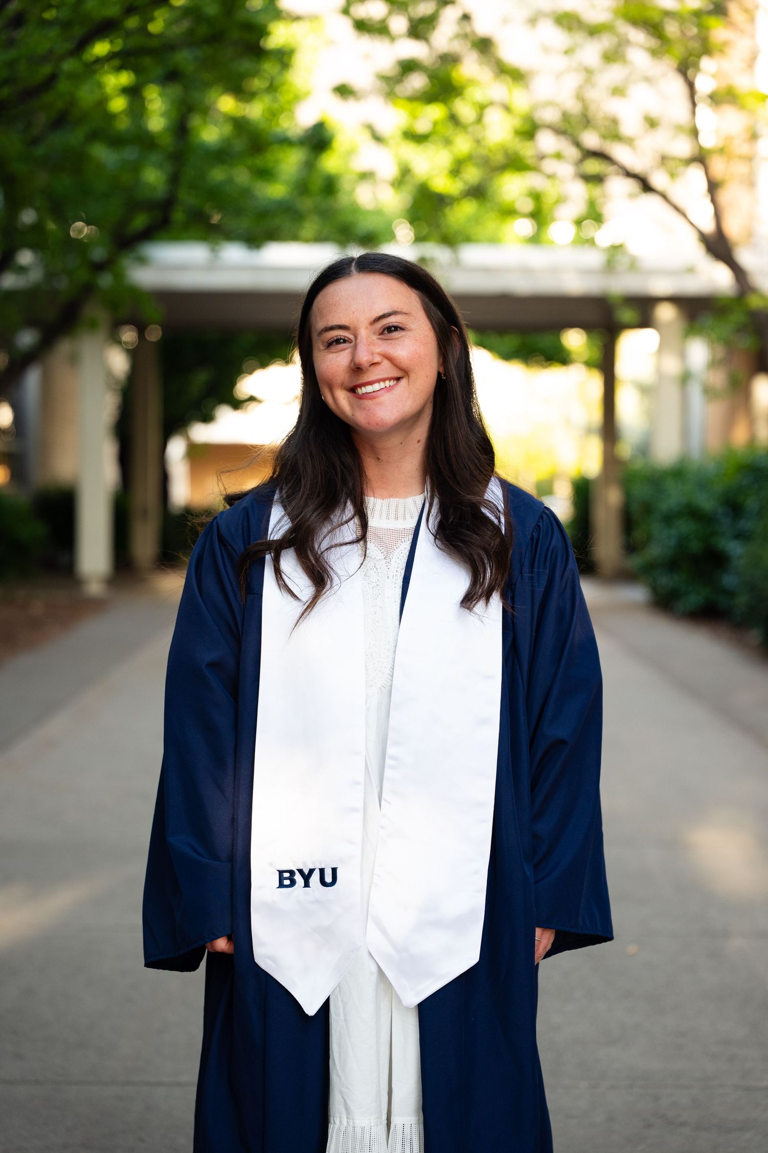 Smiling BYU graduate in navy cap and gown with white stole posing outdoors on campus.