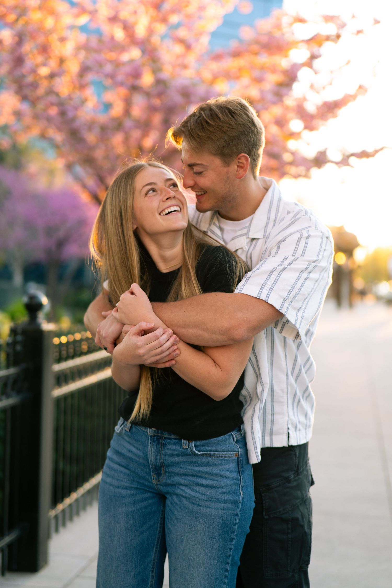 A happy couple embraces and laughs together on a sidewalk with blooming cherry blossom trees at sunset.