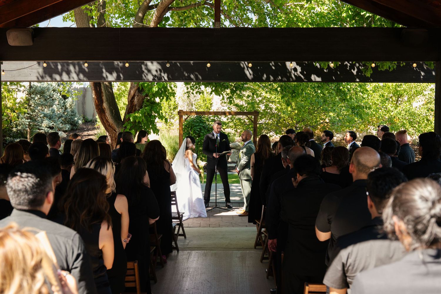 Outdoor wedding ceremony under a wooden pergola with guests seated, bride and groom at the altar surrounded by lush green trees.