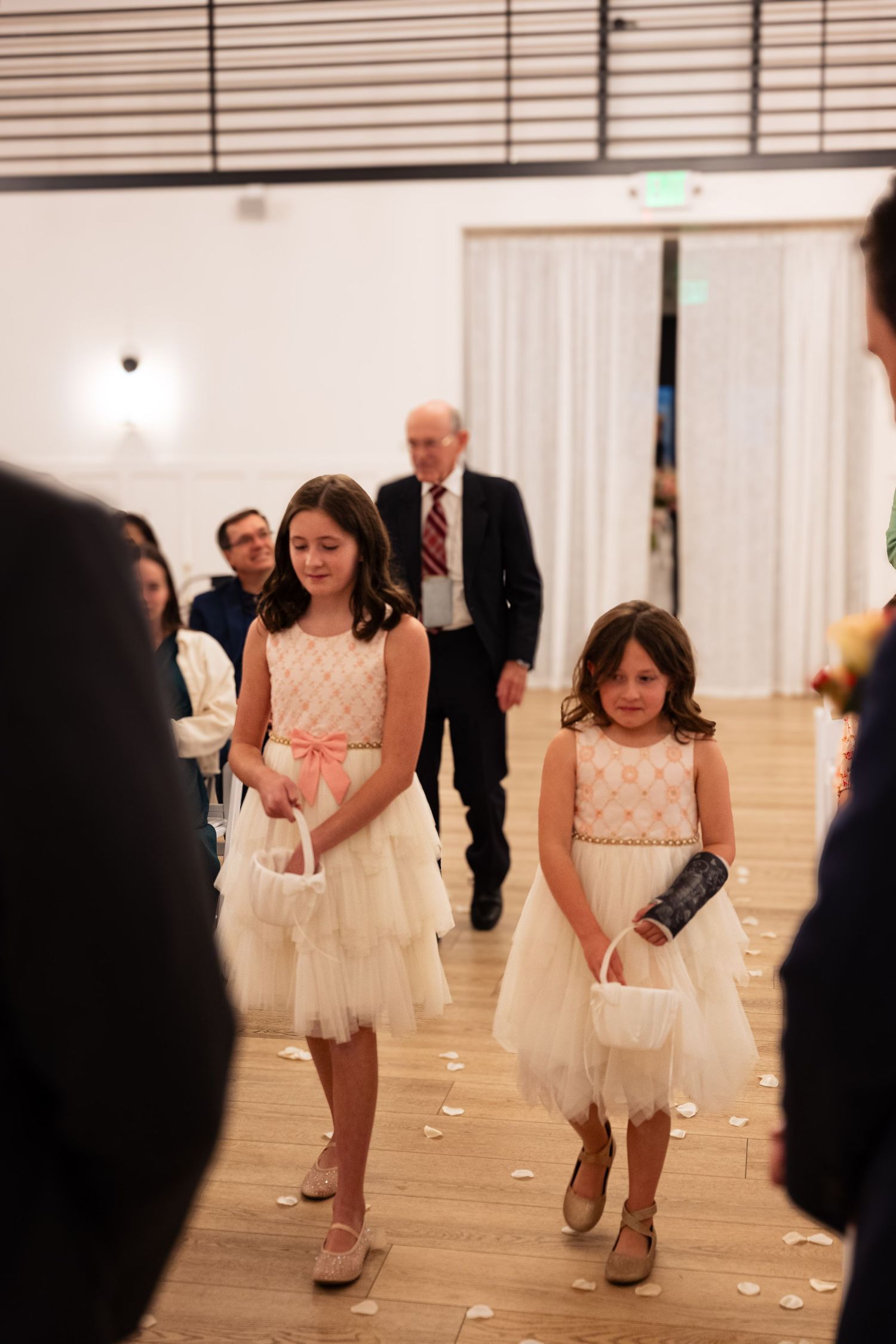 Two flower girls in white dresses walk down the aisle at an indoor wedding ceremony.