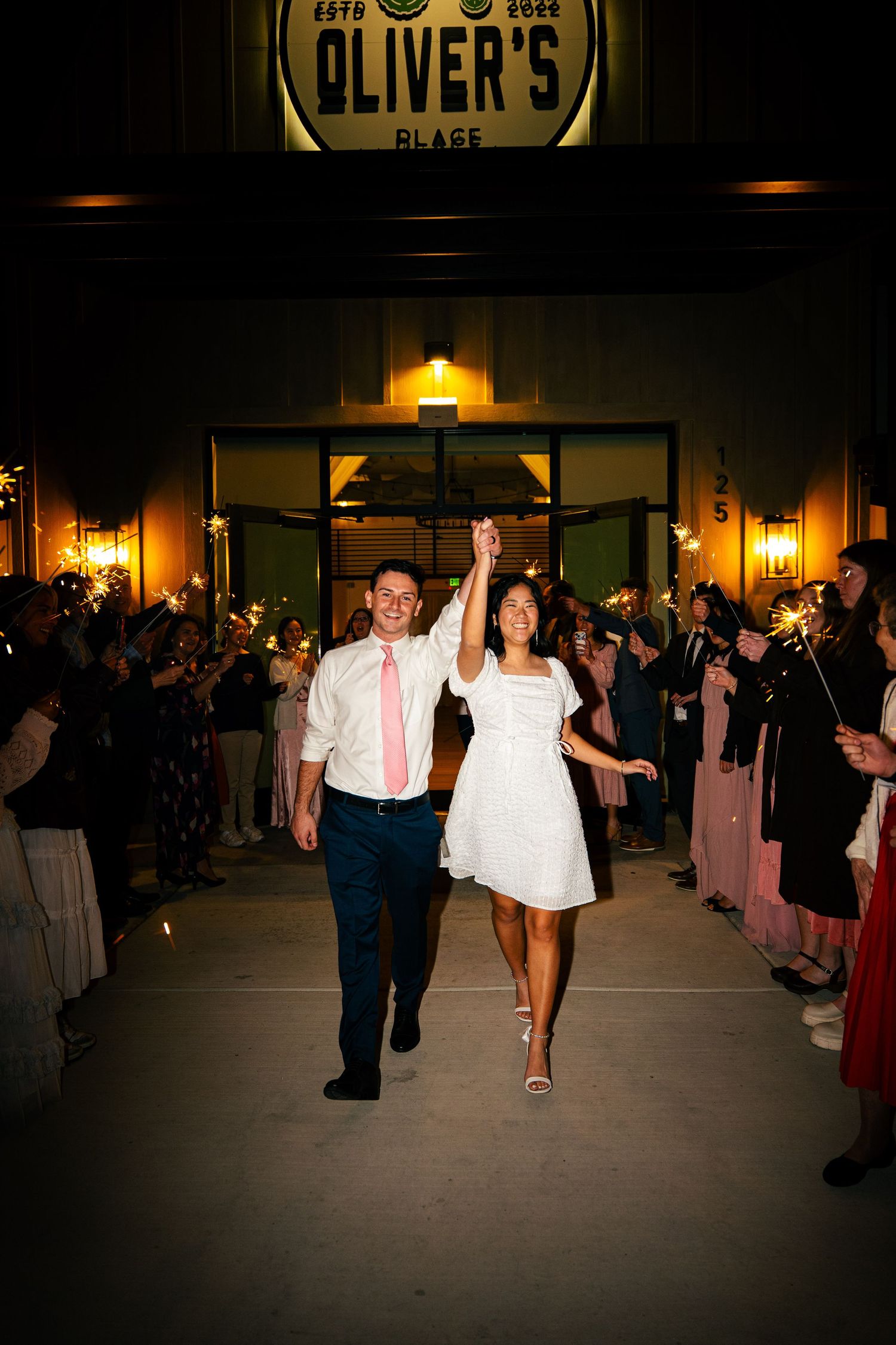 Newlyweds celebrating sparkler exit outside Oliver's Place venue, bride in white dress, groom in pink tie.
