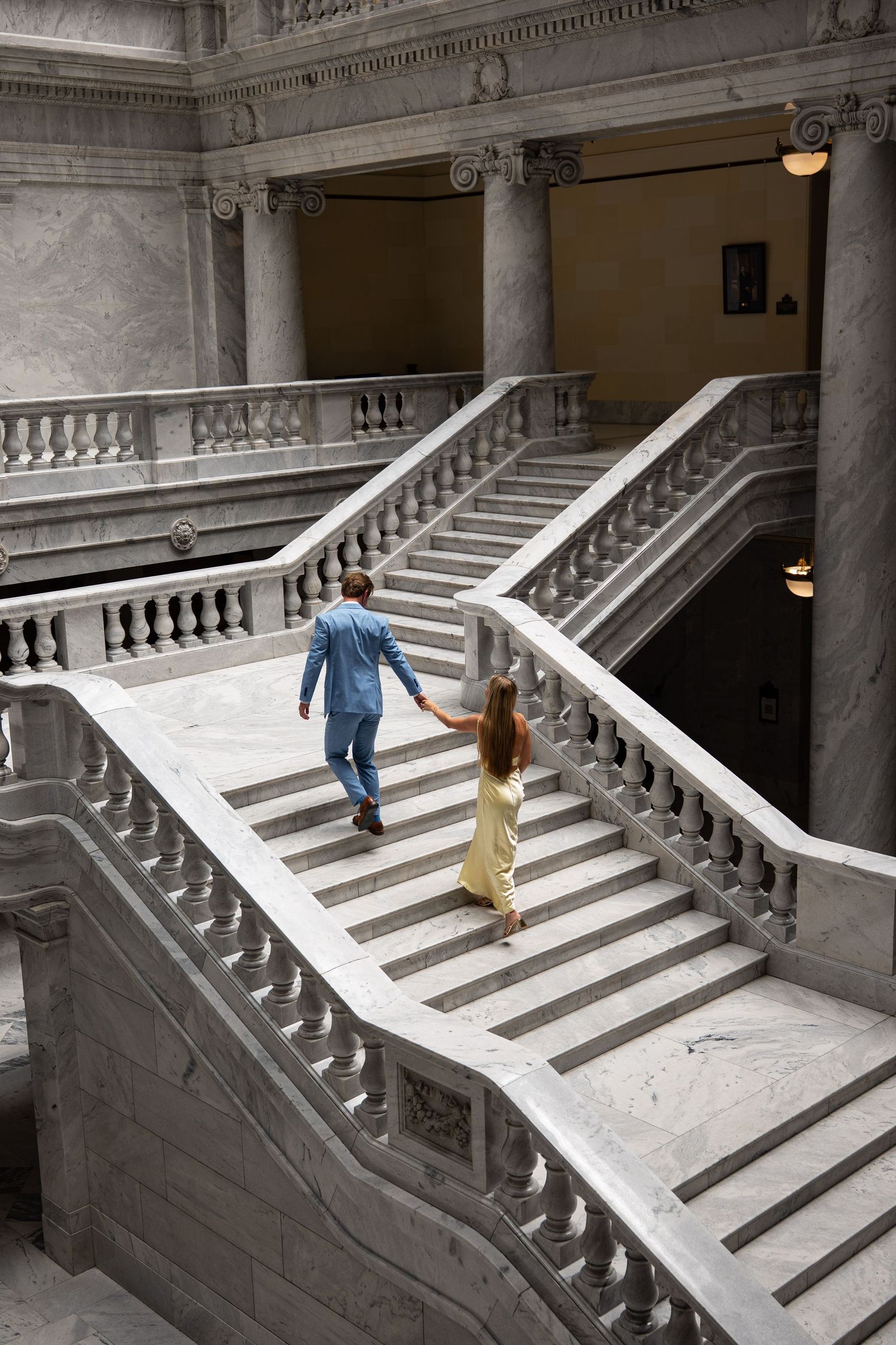 Couple holding hands walking up grand marble staircase in elegant neoclassical building interior.