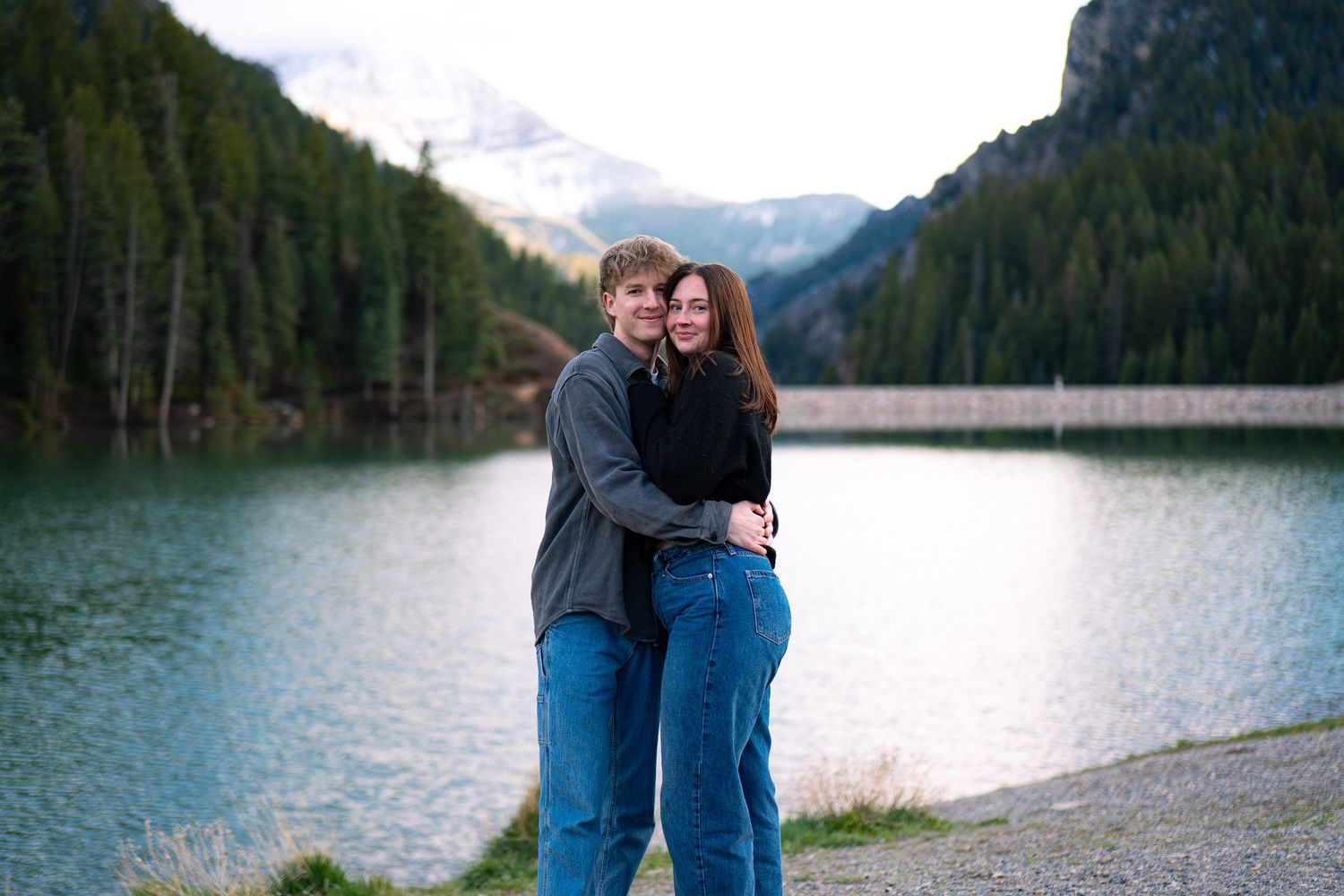 Couple embracing by a mountain lake with evergreen forest and snow-capped peaks in the background.