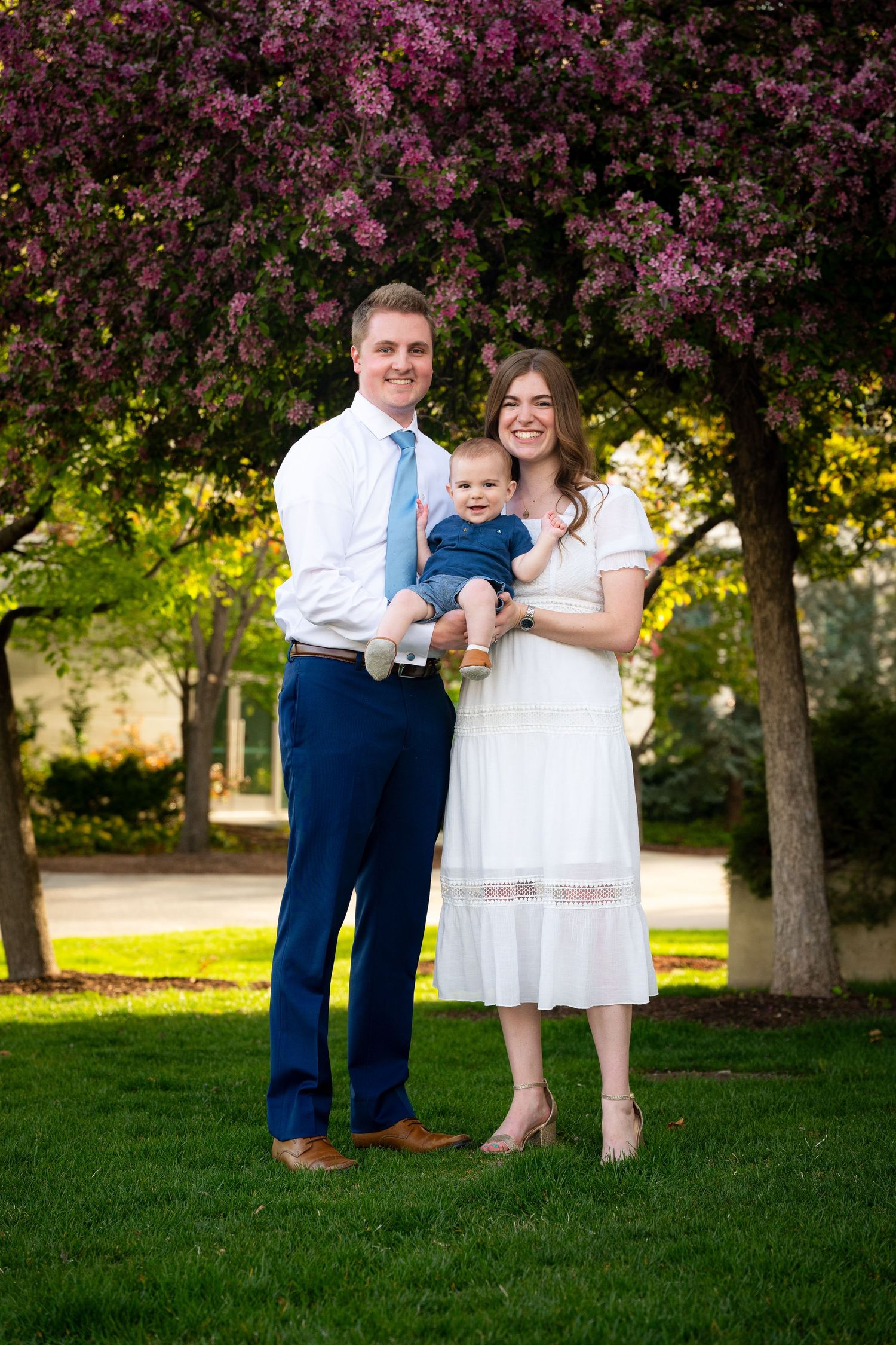 A family of three, including a young child, stands in a park with blooming trees in the background.