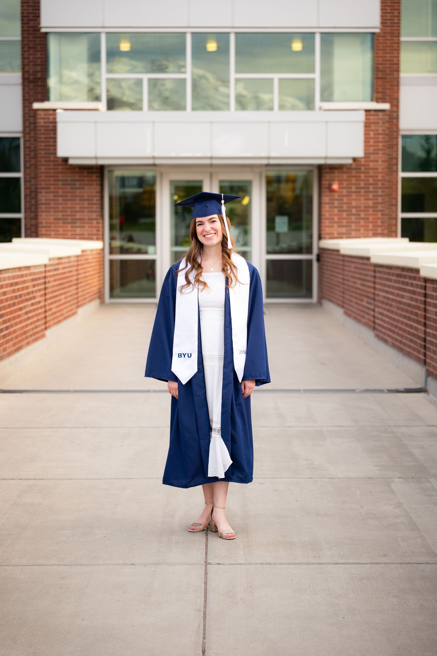 A smiling woman in a graduation gown and cap stands in front of a university building.