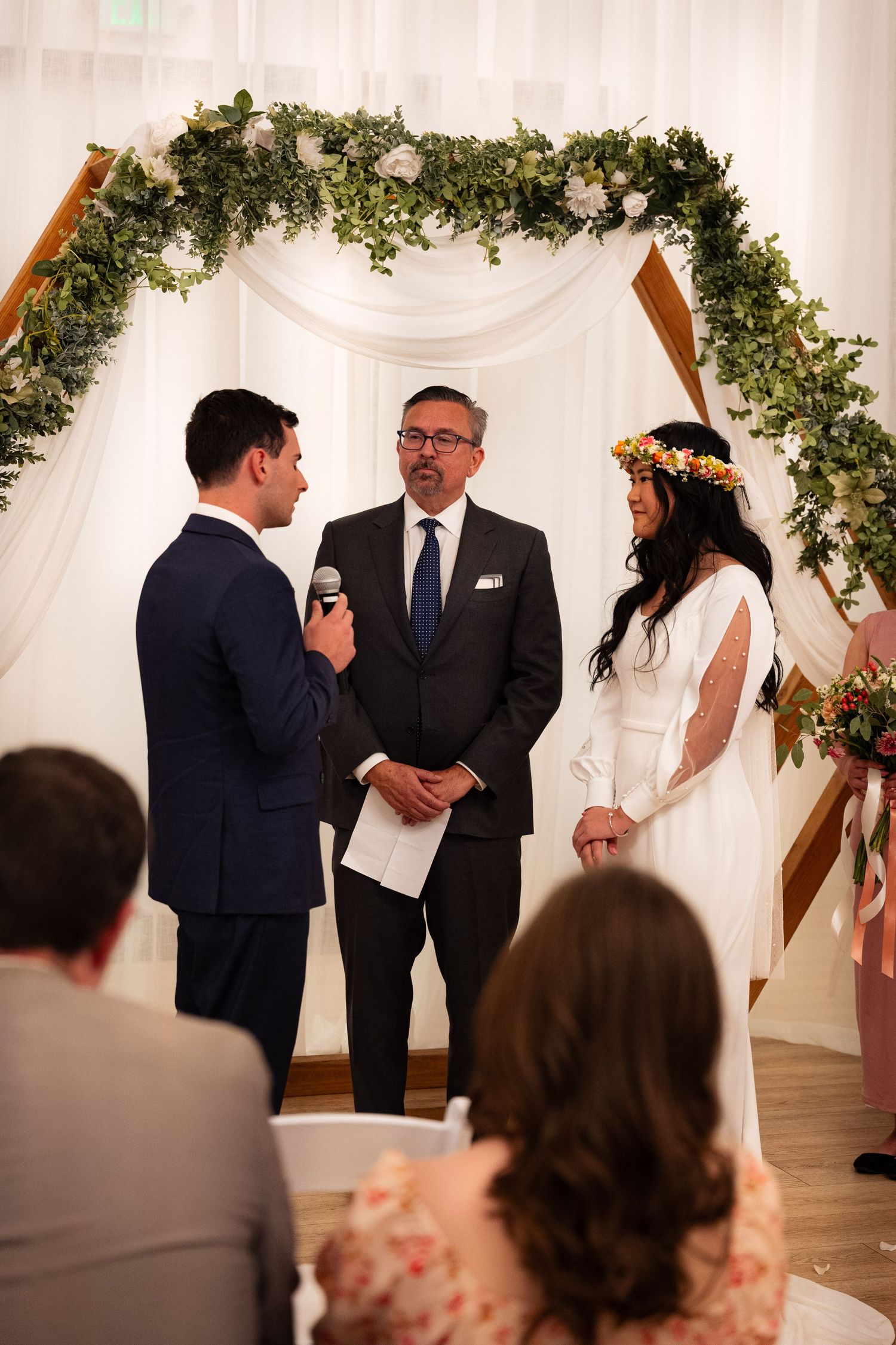 Bride and groom exchanging vows under floral arch with officiant during indoor wedding ceremony.