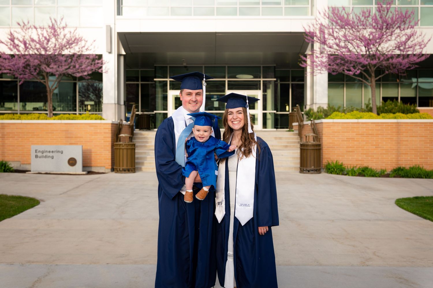 A family of three, a man, woman, and young child, stand in front of a university building.