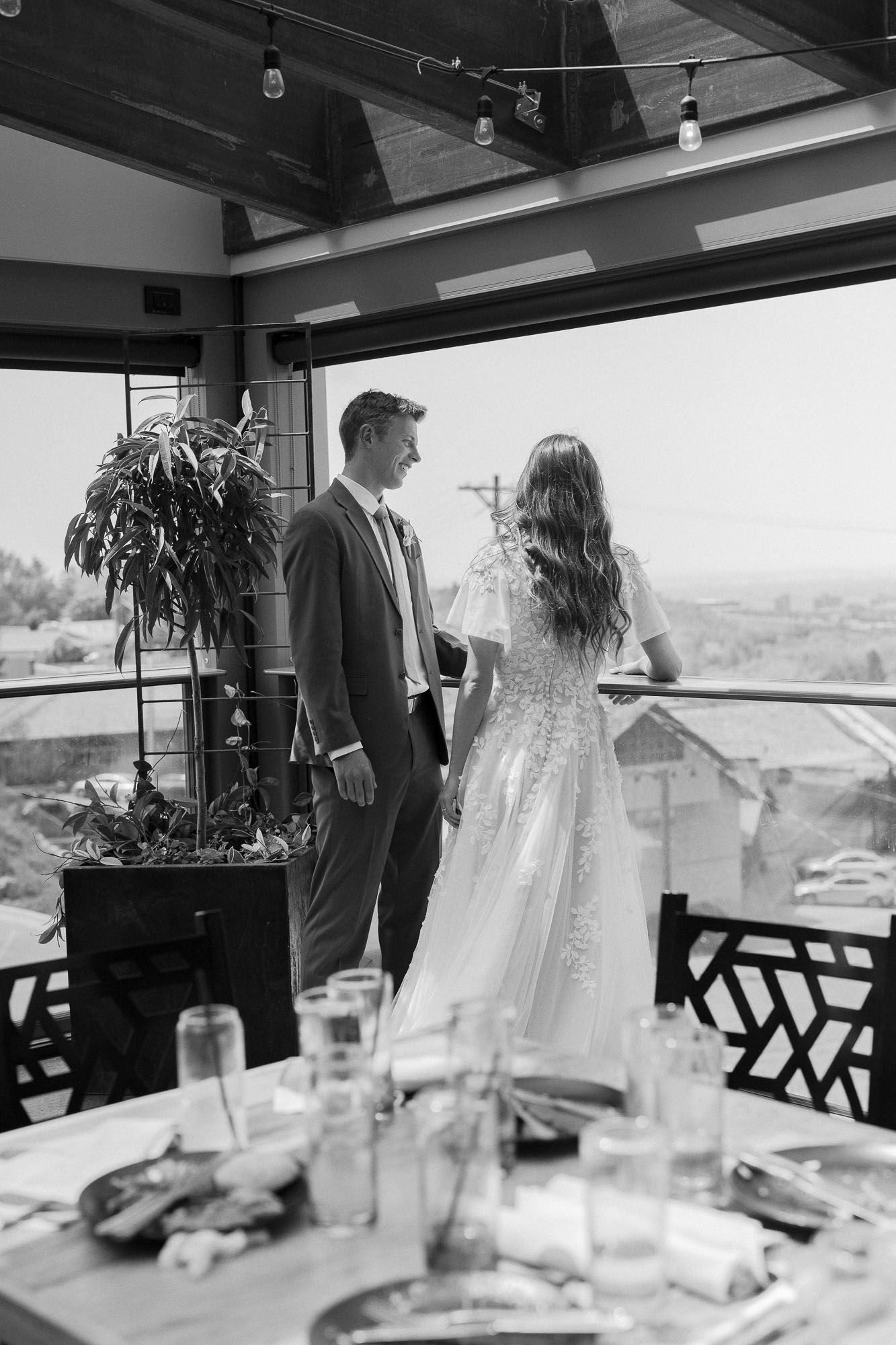Black and white wedding photo of bride in lace gown and groom in suit standing on elegant venue balcony.